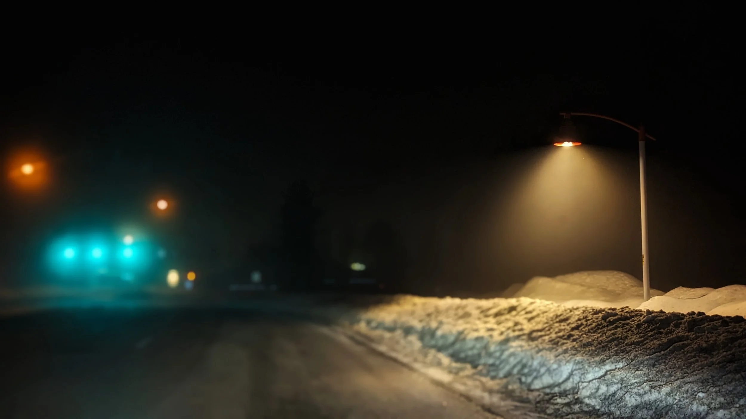 Nighttime scene of a snow-covered road with streetlights illuminating the area, and traffic lights visible in the distance.