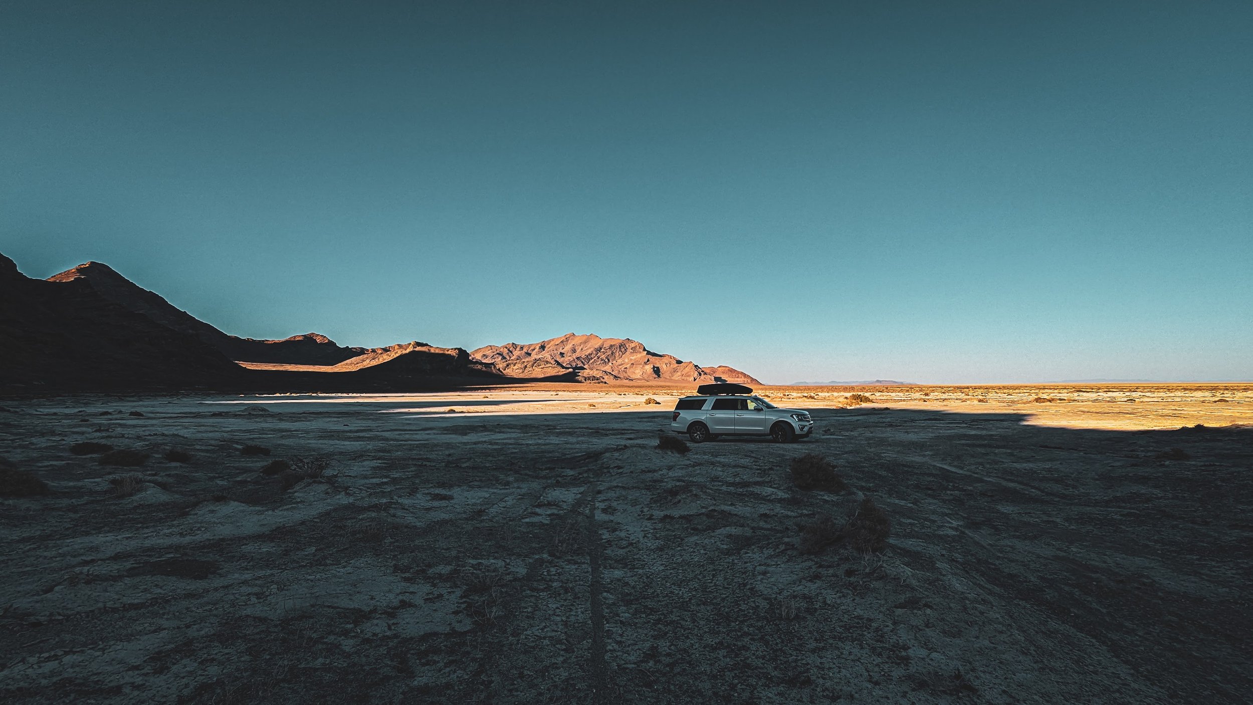 A white SUV parked in a desert landscape with mountains in the background and a clear blue sky.