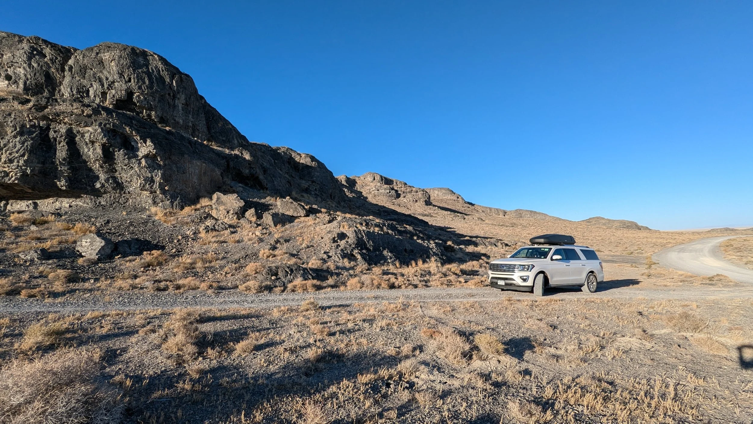 A white SUV with a rooftop cargo box parked on a dirt road in a mountainous desert landscape with rocky hills and clear blue sky.