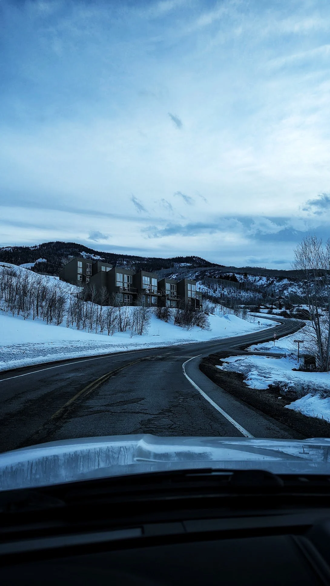 A winding road runs through a snowy landscape with modern buildings on a hillside, under a cloudy sky.