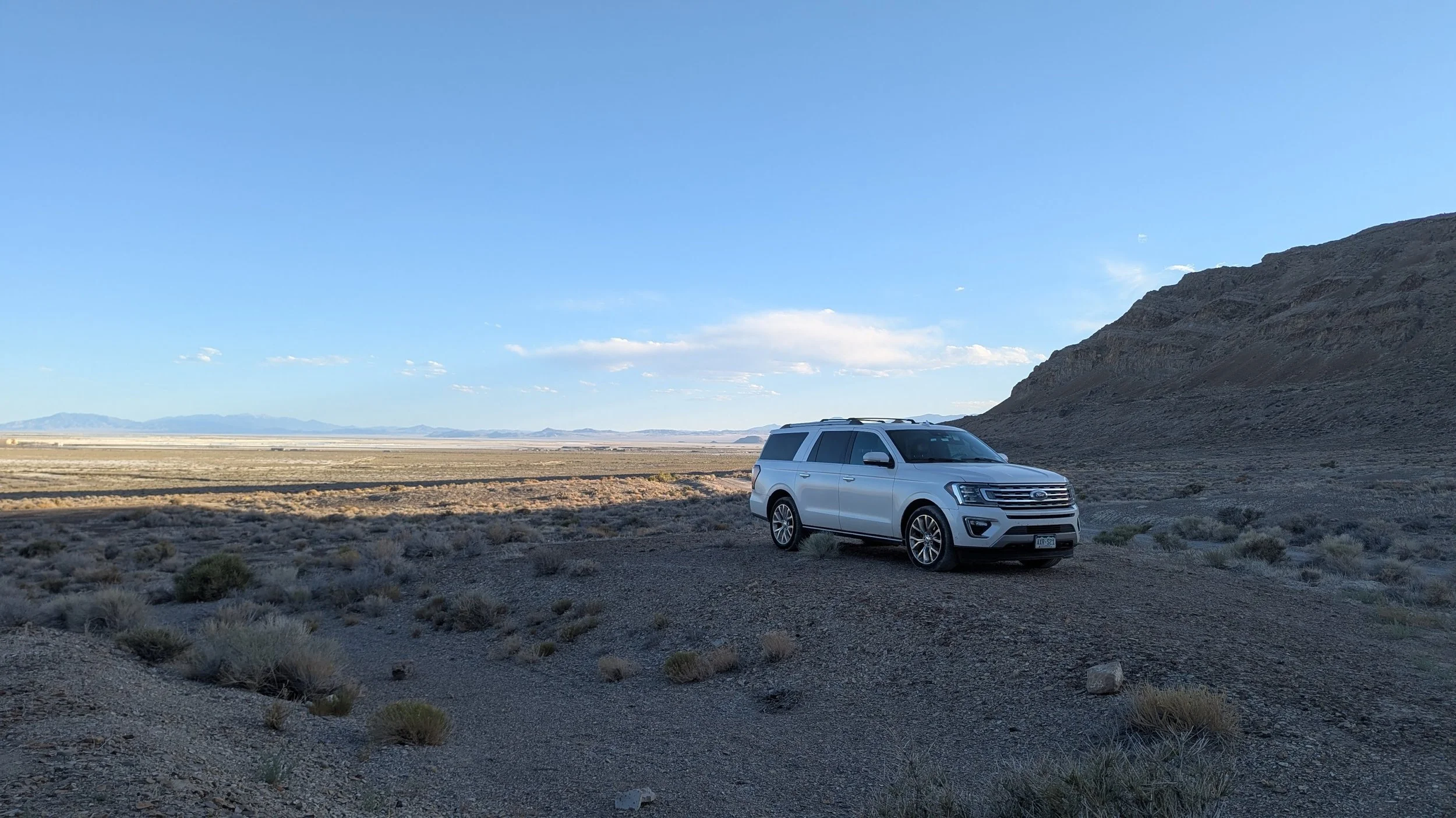 A white SUV parked on a dirt patch in a desert landscape with sparse vegetation, mountains in the background, and a clear blue sky.
