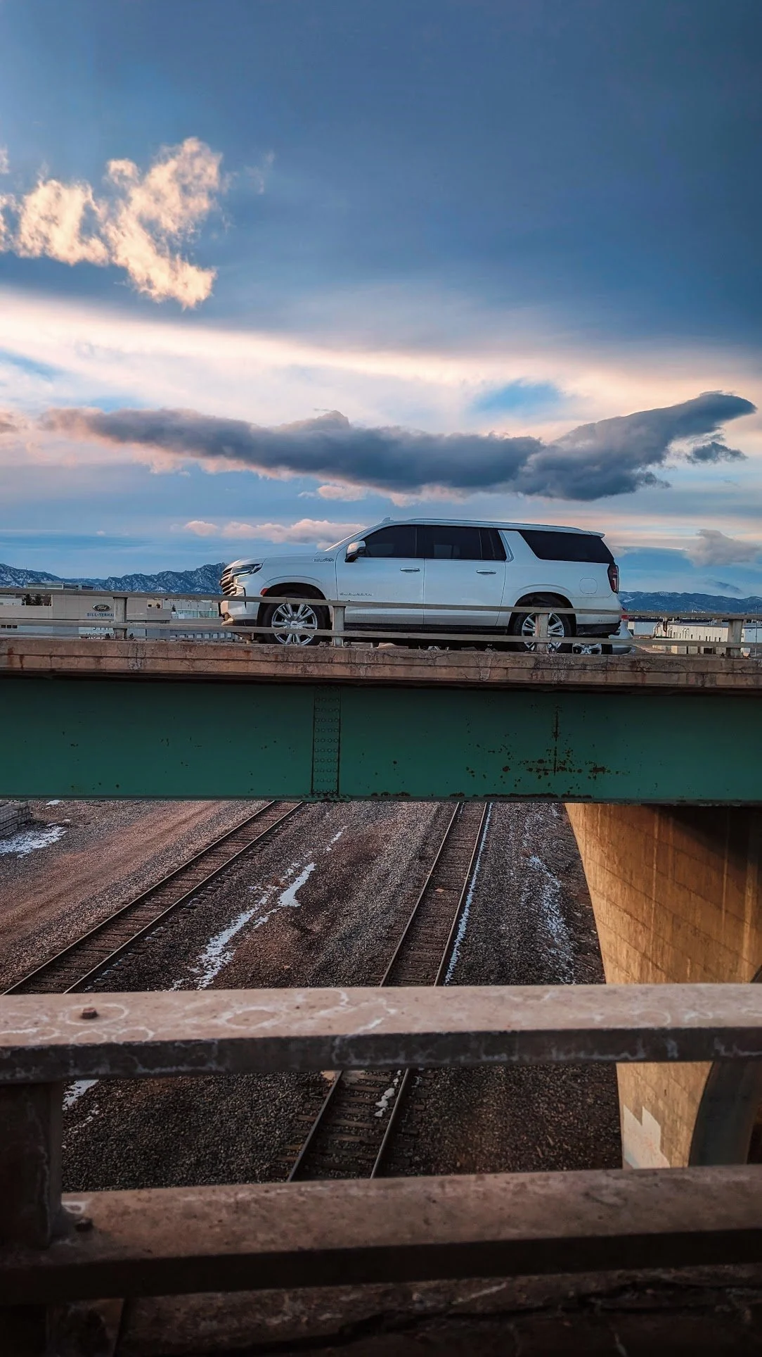 A white SUV parked on top of a green bridge over railway tracks during sunset with clouds and mountains in the background.