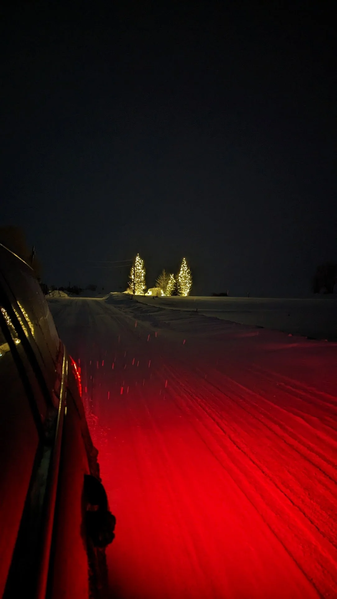 Nighttime snowy road with holiday lights on trees in the distance, viewed from the side of a vehicle with red taillights reflected on the snow. Dreamscape. Fairytale Destinations. Mountain Trip. Ski season. Steamboat Springs.