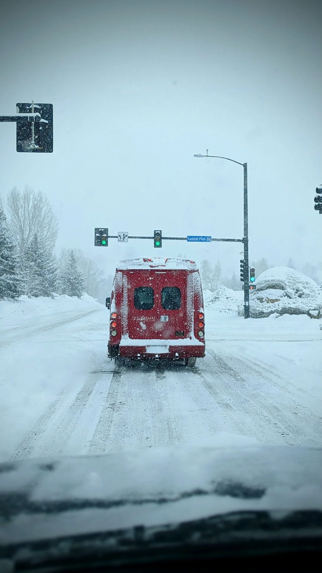 Snow-covered road with a red vehicle, traffic lights, and snow-covered trees and rocks on the sides, in a winter setting.