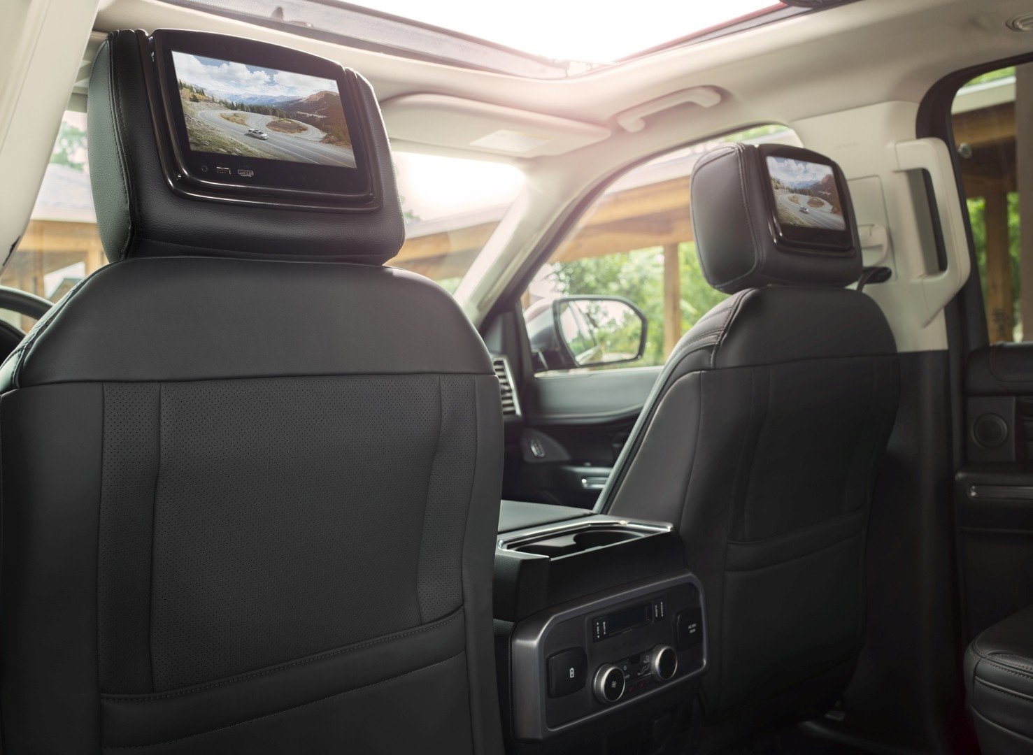 Interior of a vehicle showing the back of the front seats with headrest screens displaying a mountain landscape scene, and a control panel with climate controls at the rear of the center console.