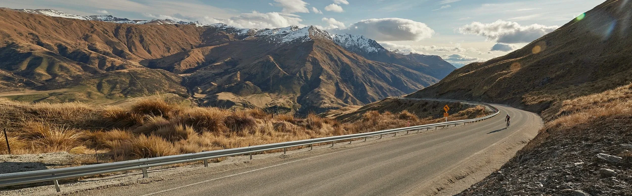 a cyclist on a curved road riding uphill with snow capped mountains in the background