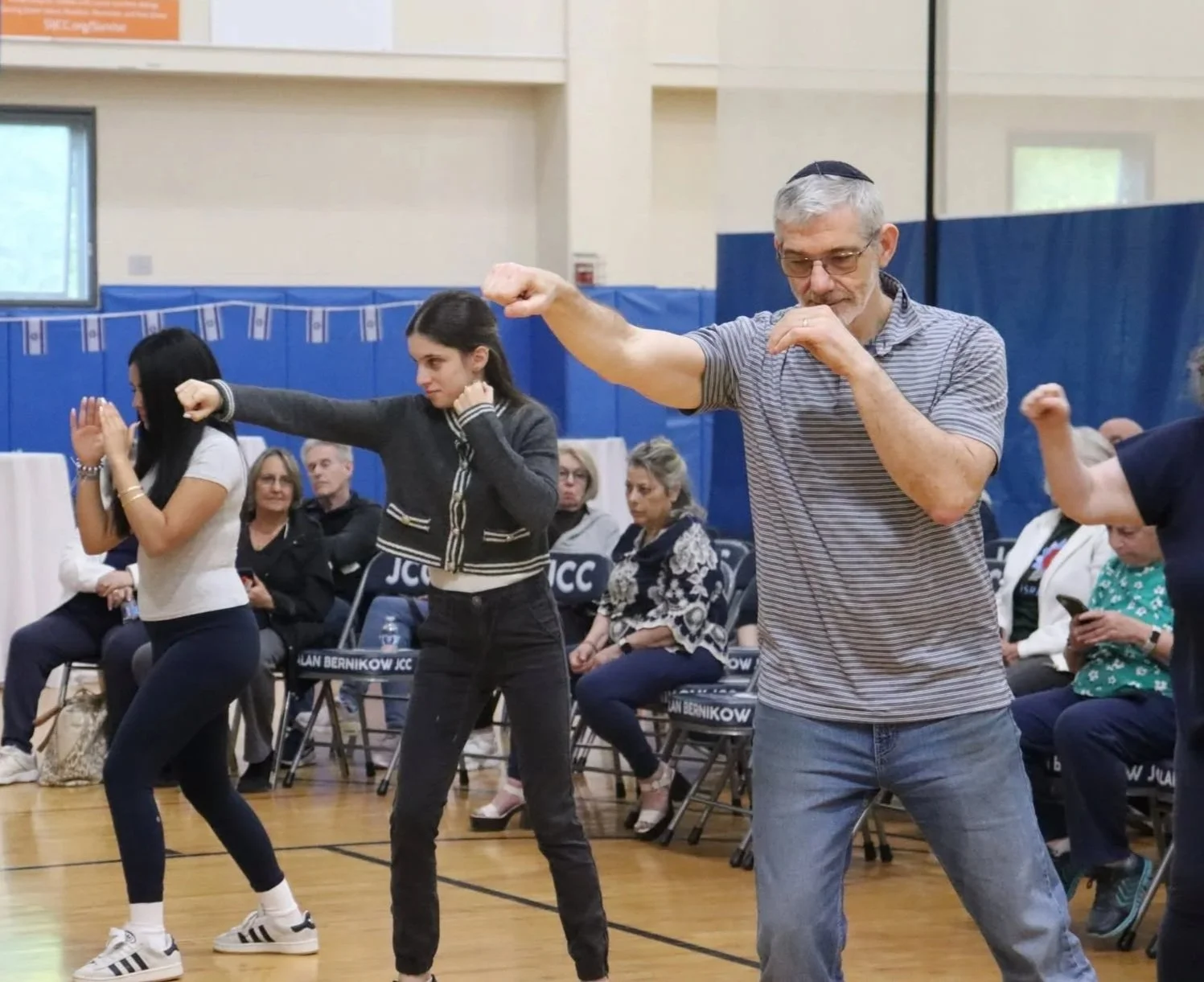 Group of people participating in a dance or aerobic class indoors, with a man leading, all in a gymnasium with seating and blue walls.