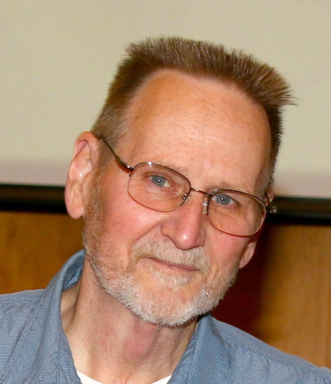 Close-up of an older man with glasses, short hair, and a beard, wearing a blue shirt.