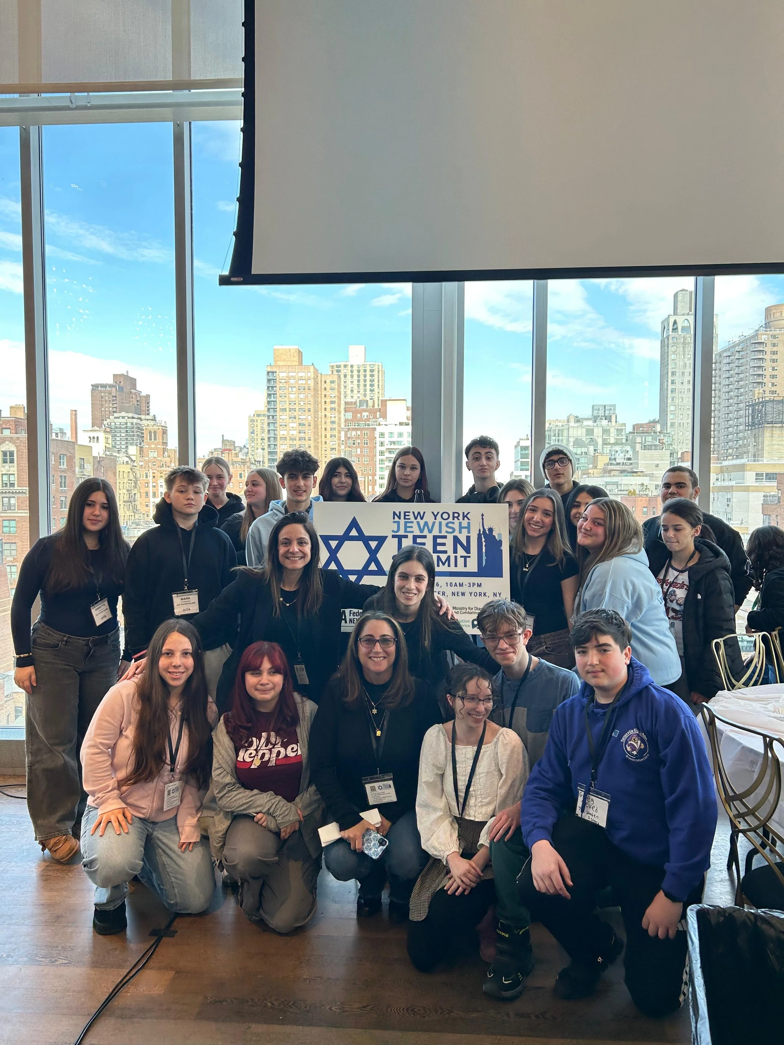 Group of teenagers and adults at the New York Jewish Teen Summit, standing indoors near large windows with a cityscape background.
