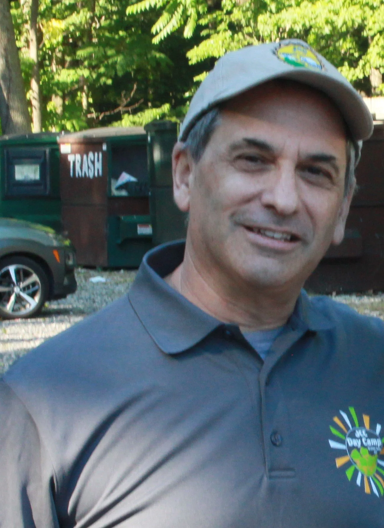 A man wearing a gray polo shirt and a beige cap, smiling outdoors with green trees and a trash bin in the background.