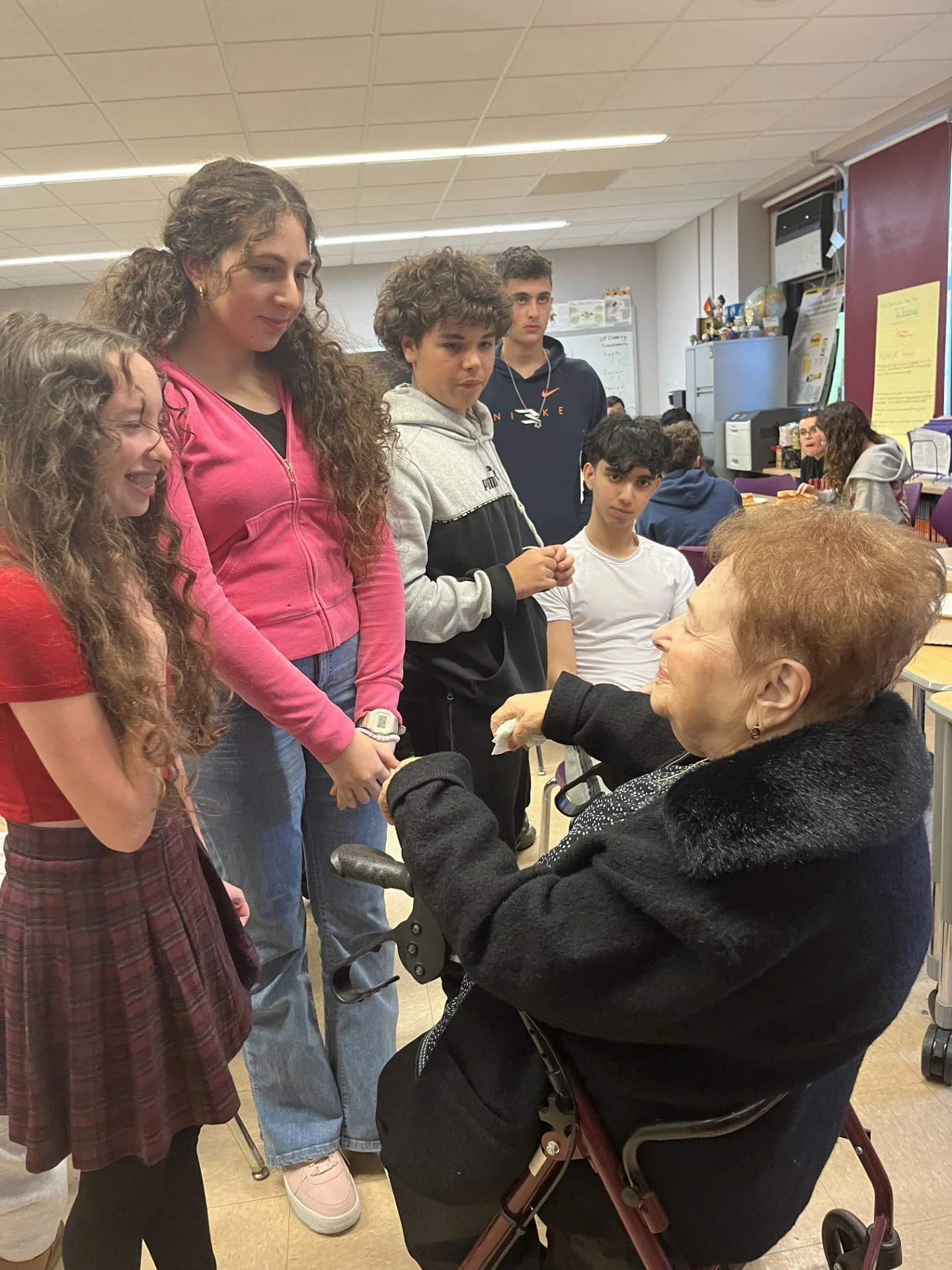A group of five teenagers, four standing and one sitting in a wheelchair, interacting with an elderly woman in a classroom or community center setting.