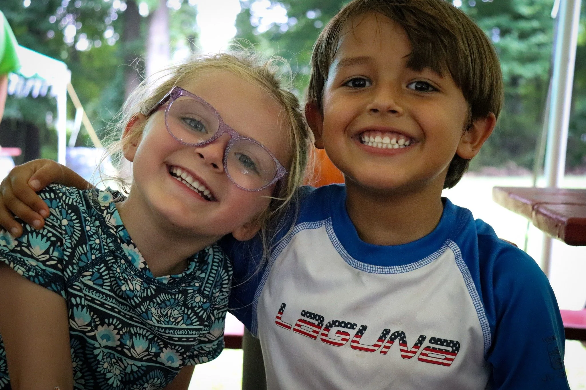 Two children, a girl with blonde hair and glasses and a boy with dark hair, smiling and hugging at an outdoor event in a park.