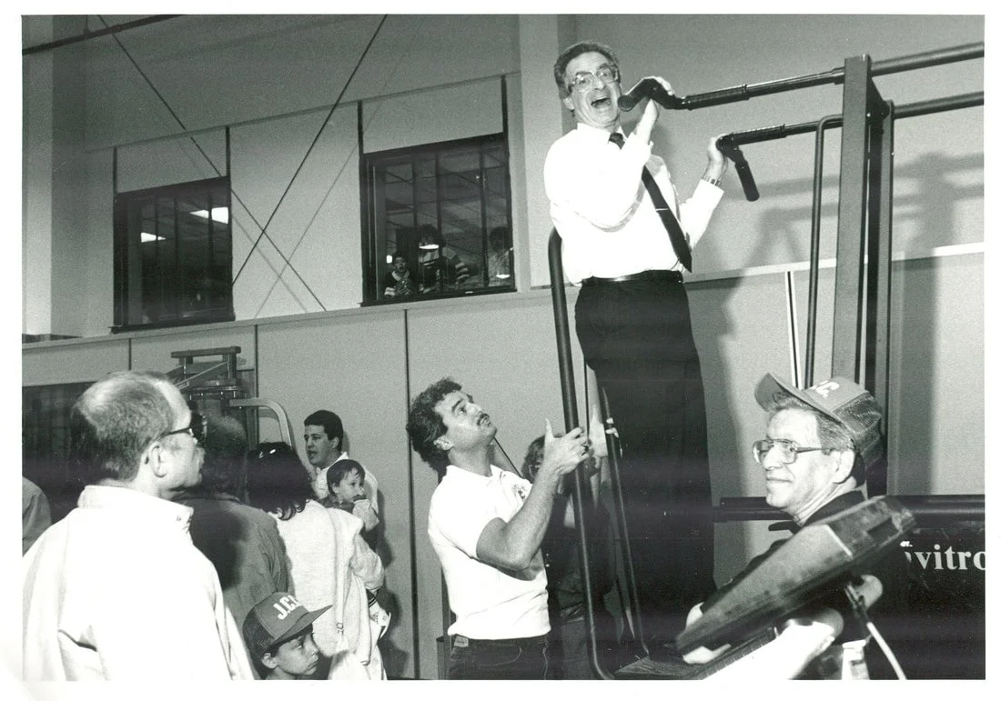 A man standing on a platform, smiling and speaking into a microphone connected to a pipe, with a crowd watching and a woman holding a child, all indoors.