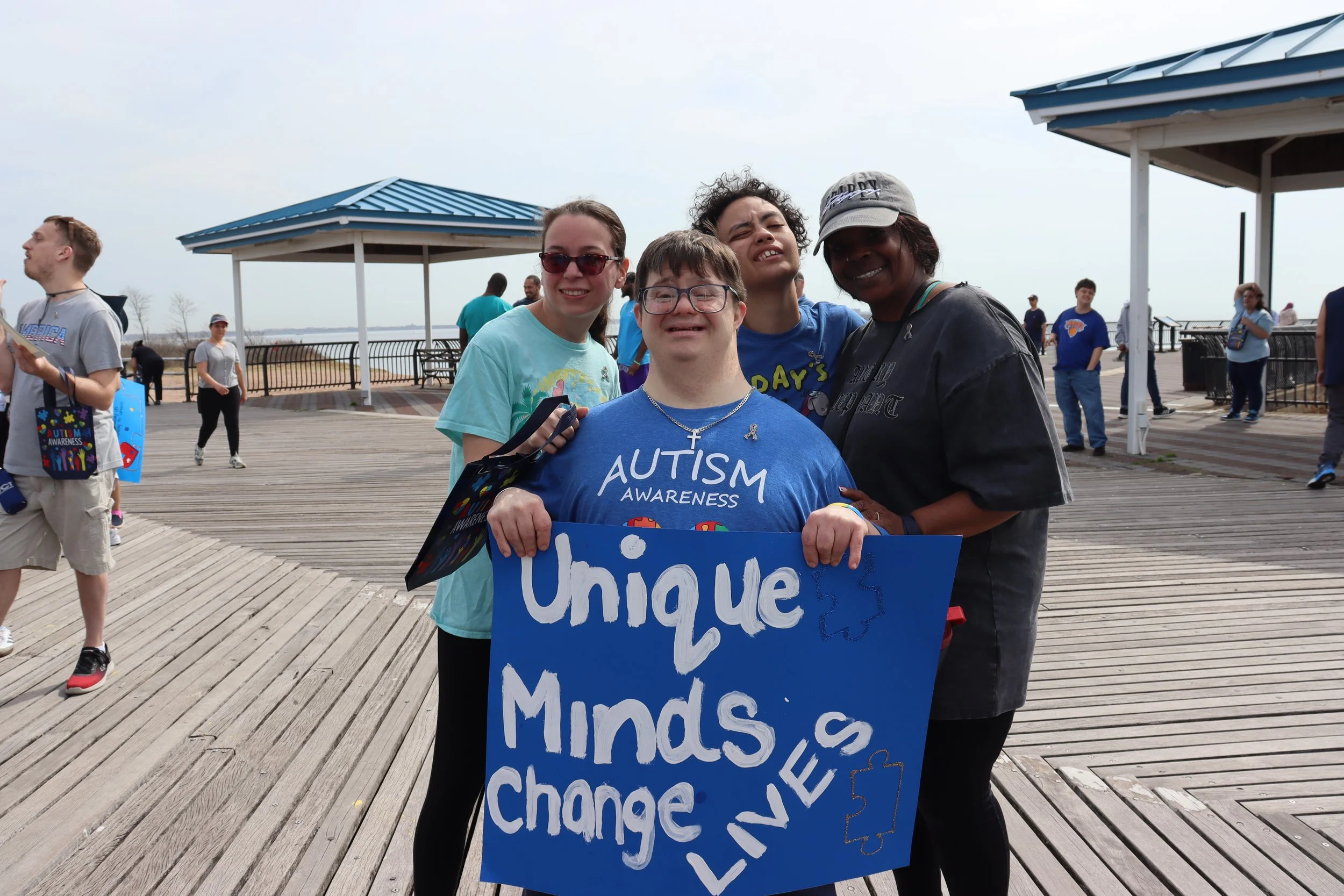 A group of people participating in an autism awareness event on a wooden boardwalk outside. There are five people in the foreground, smiling and holding a blue sign that reads 'Unique, M.
