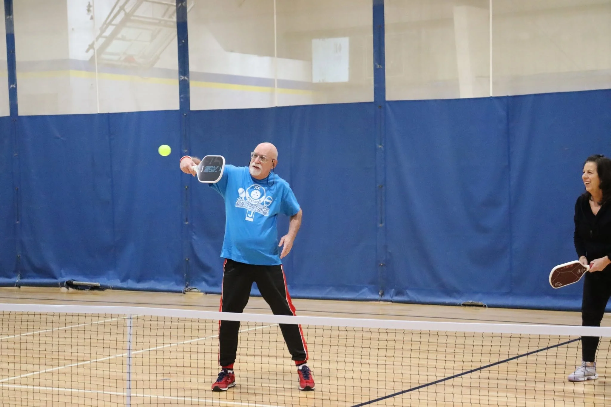 An elderly man with glasses playing pickleball on an indoor court, using a paddle and a green ball, with a woman holding a paddle smiling nearby.