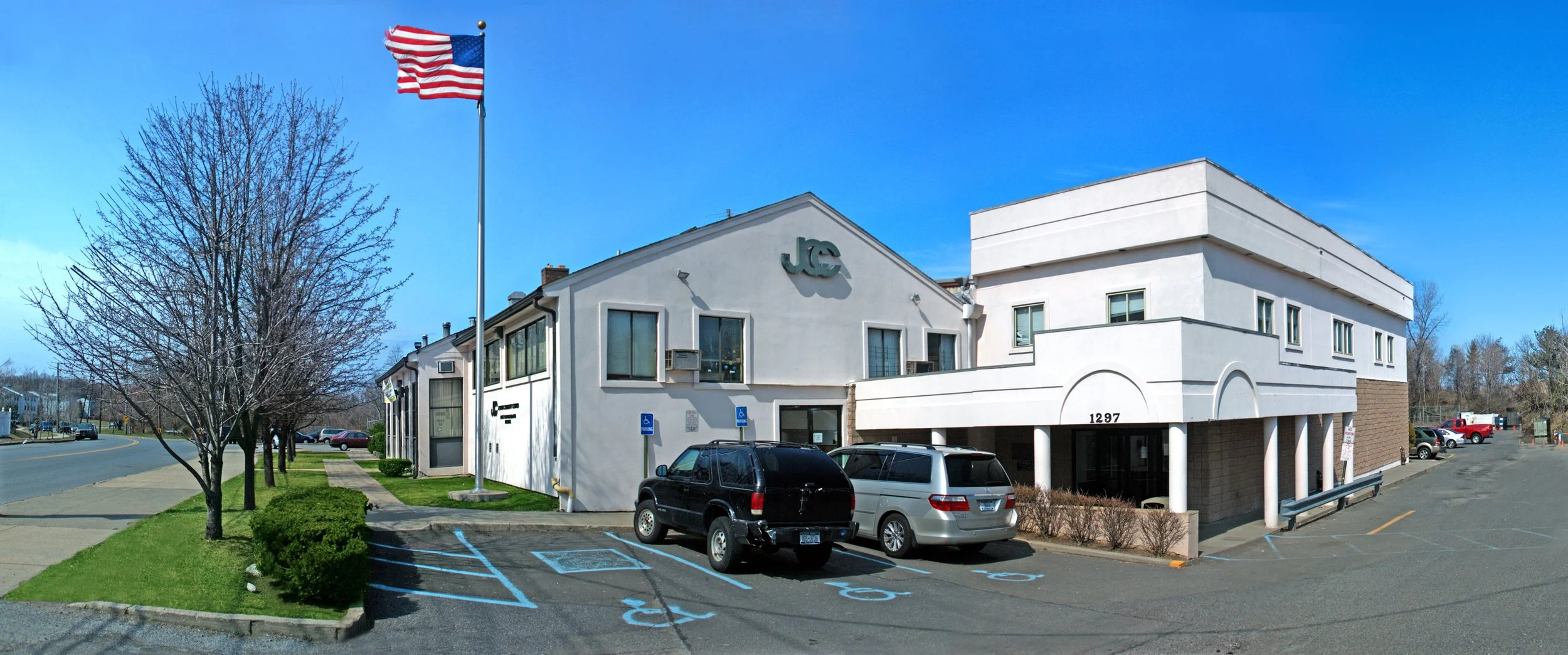 A two-story white commercial building with the letters 'J C' on the front, parking lot with cars, and an American flag flying in front of a leafless tree against a bright blue sky.