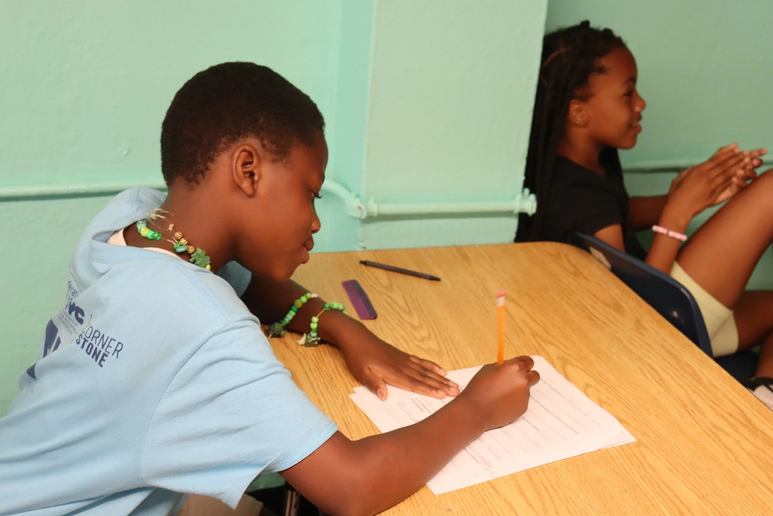 A girl with short hair, wearing a light blue T-shirt and beaded jewelry, is sitting at a wooden table, writing in a notebook with a pencil. Another girl with long braids and a dark shirt is sitting beside her, engaged in prayer or meditation with hands clasped. The room has a light green wall in the background.