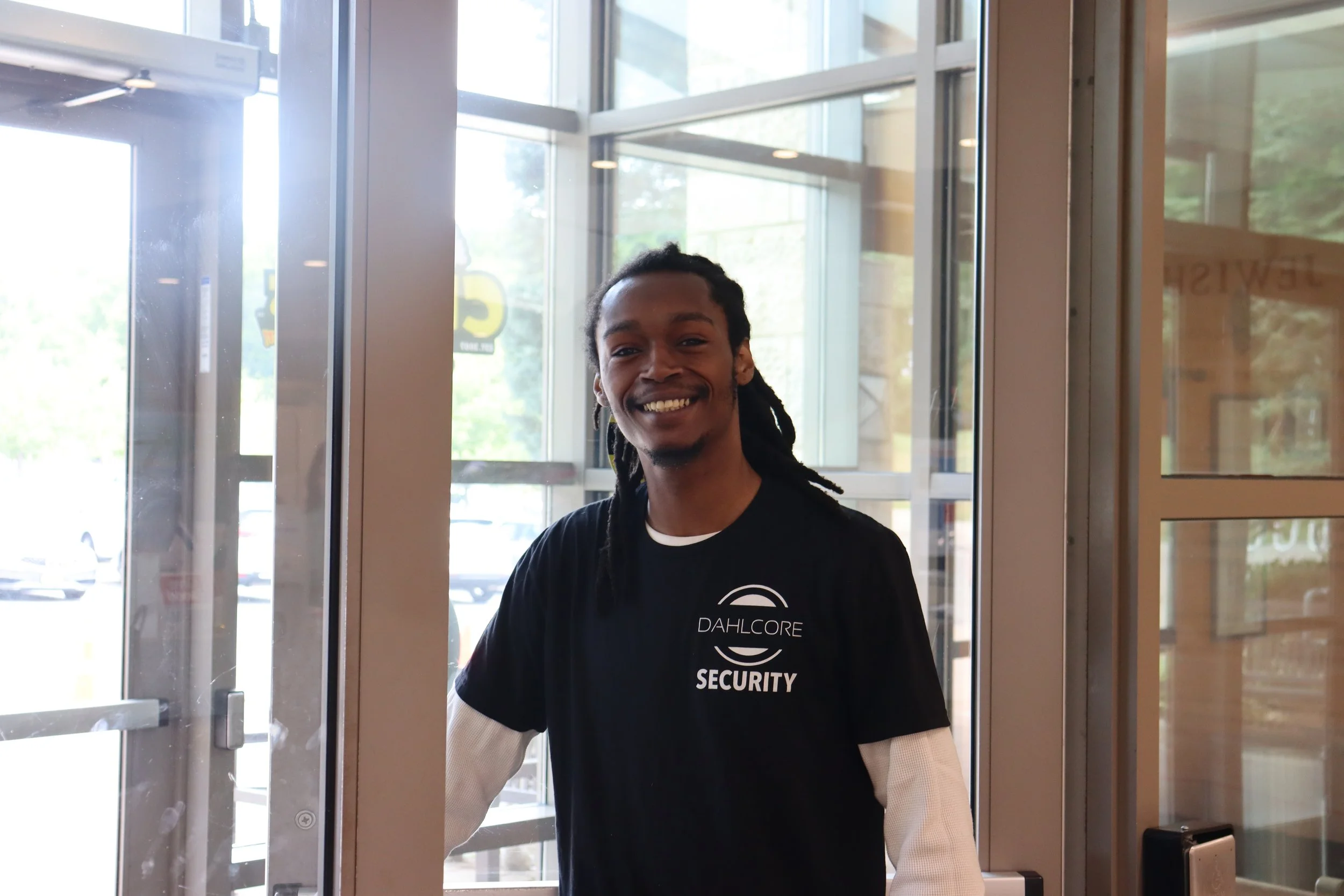 A smiling man with long dreadlocks and a goatee standing at the entrance of a building, wearing a black security T-shirt with white long sleeves underneath, near glass doors with metal framing.