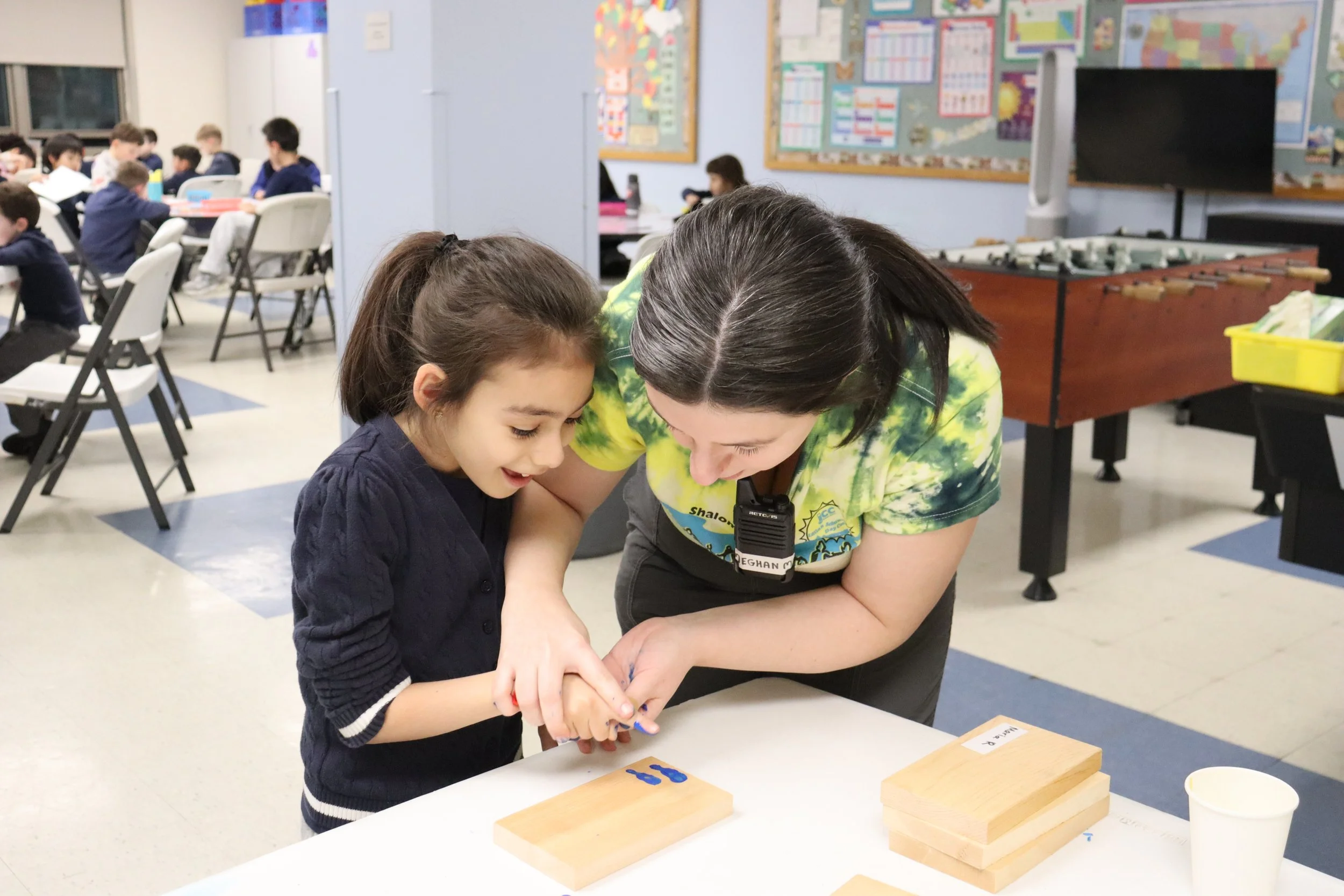 A young girl and an adult woman working together, examining something on the table with wooden blocks and a small block with the word 'MORE' on it in a classroom setting.