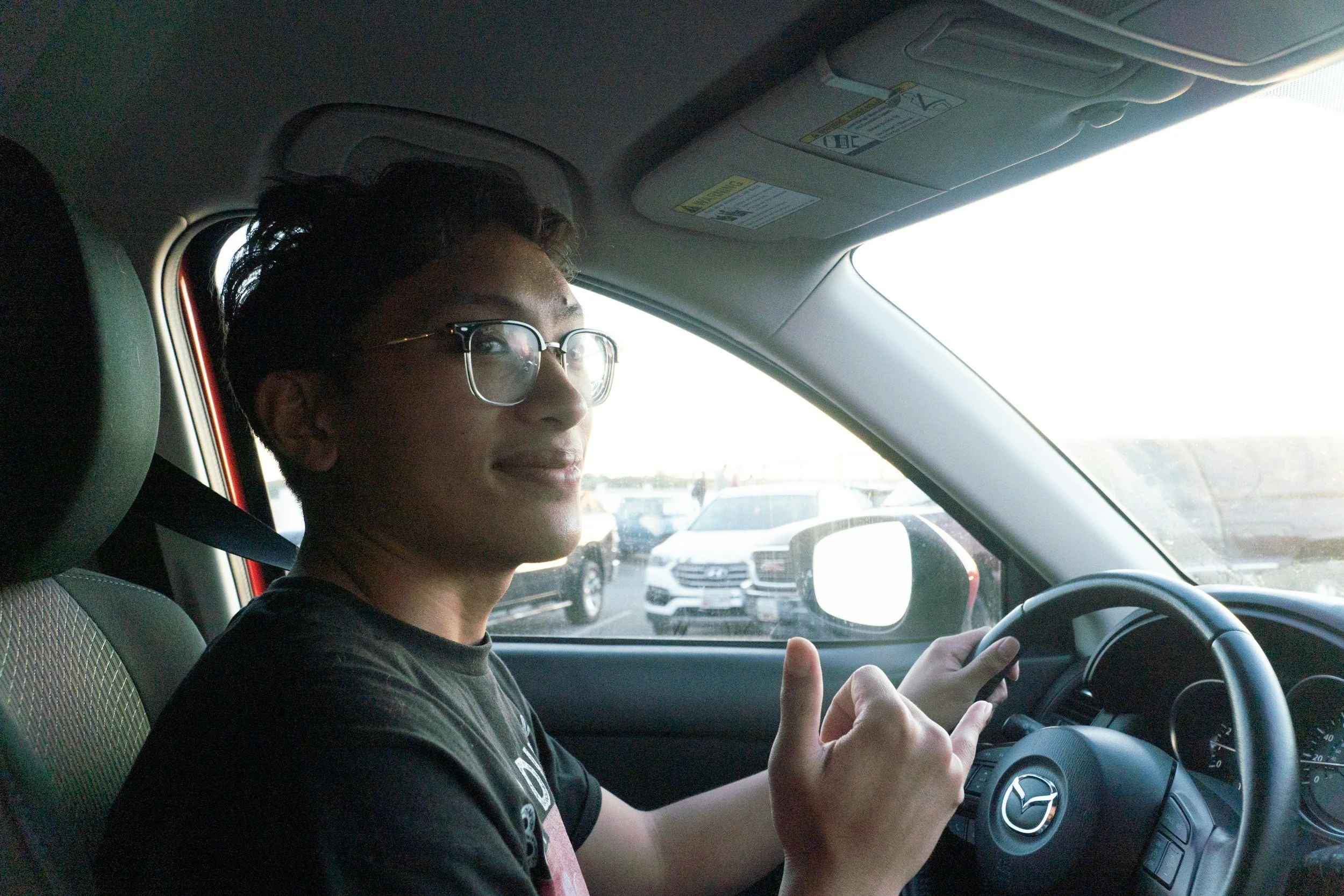 A young man with glasses sitting in the driver's seat of a car, giving a thumbs-up gesture, with vehicles visible through the window.