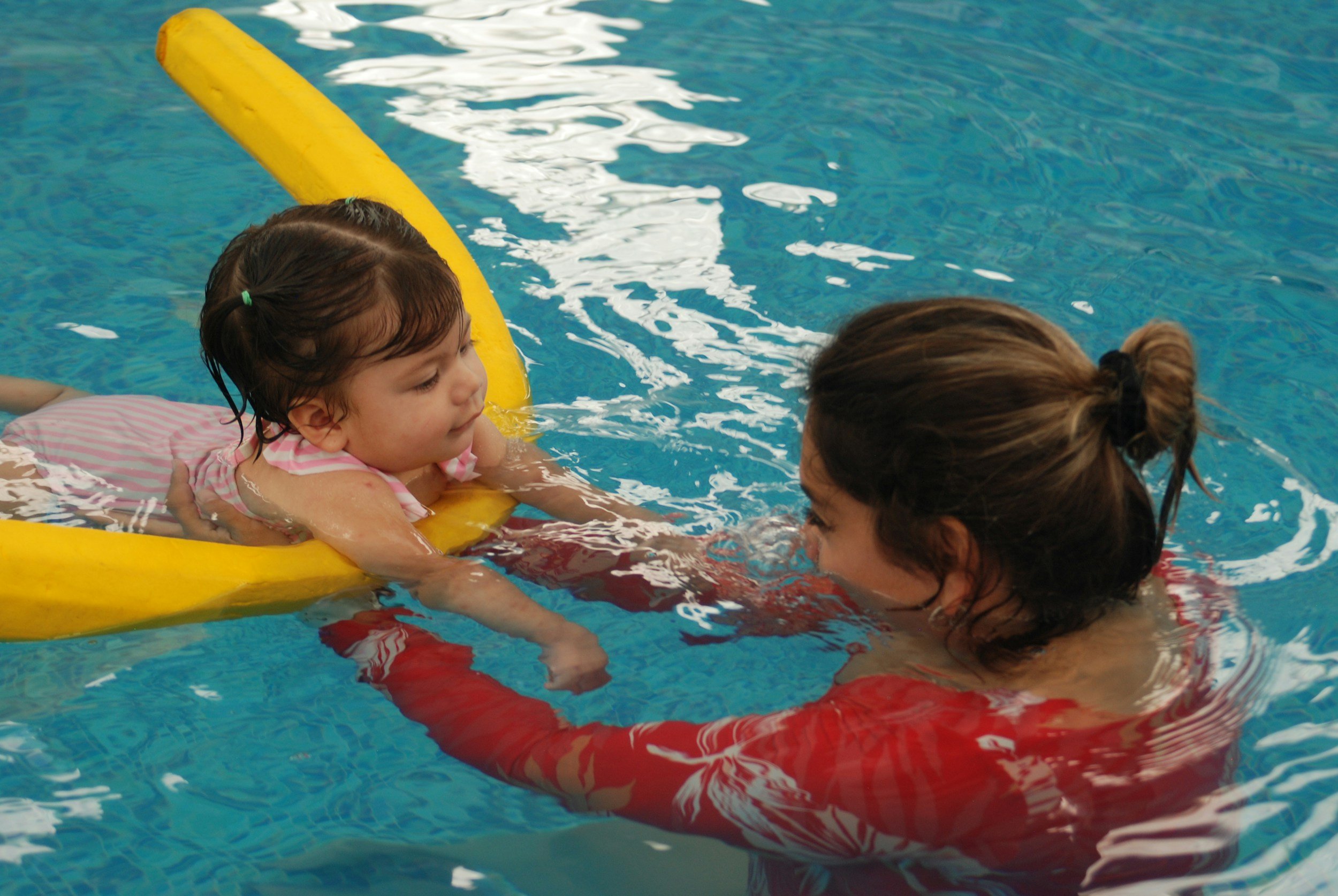 A young girl practicing swimming with an instructor in a pool, using a yellow floatation device and holding onto a red lane divider.