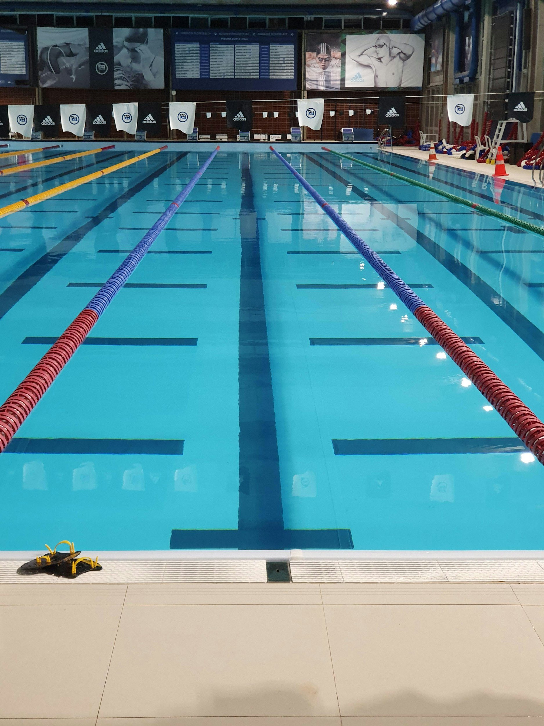 Indoor swimming pool with empty lanes, a pair of water shoes at the foreground, and folding chairs and orange traffic cones along the poolside. There are advertisements and a schedule on the wall behind the pool.
