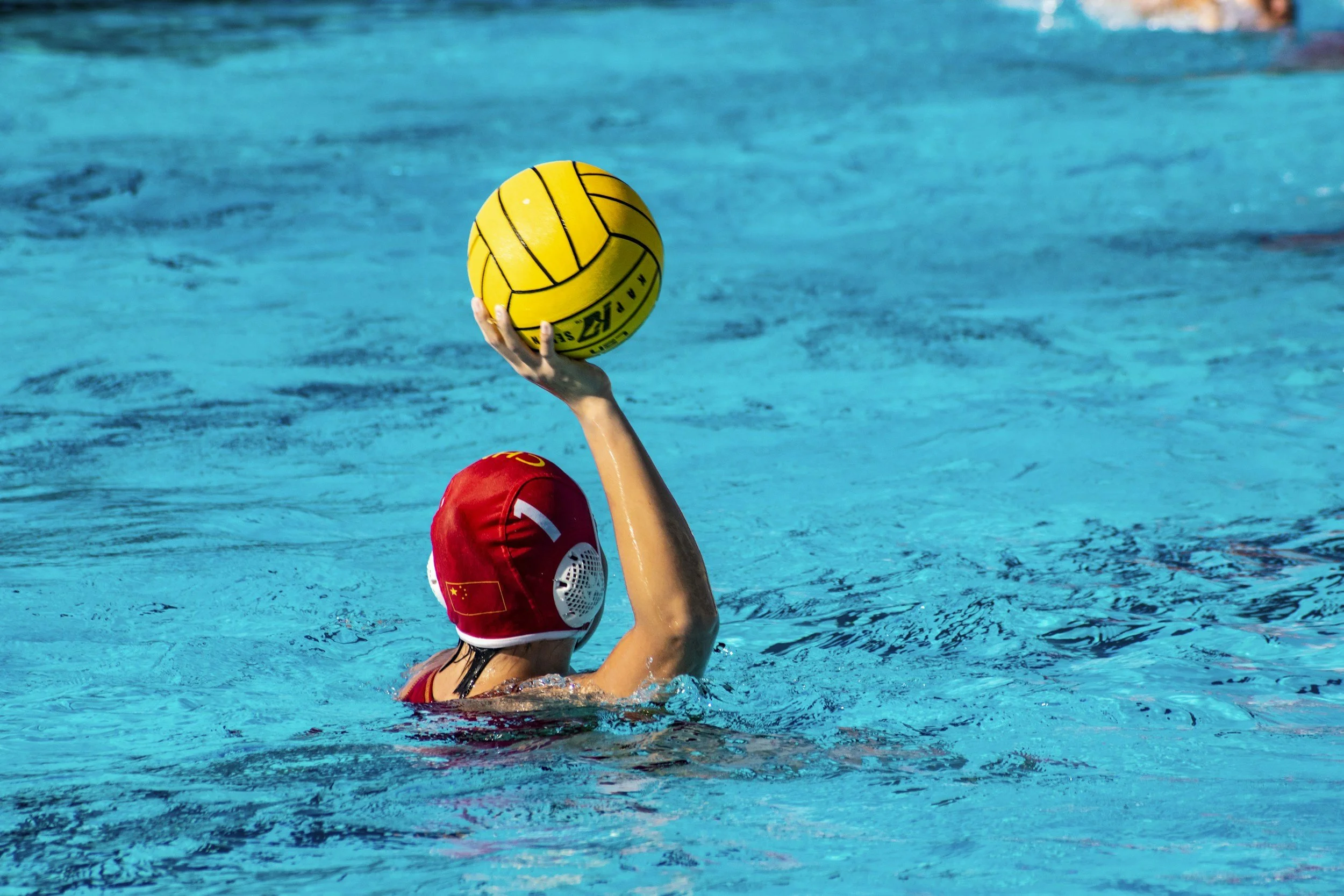A water polo player wearing a red cap with a Chinese flag is reaching up to catch or pass a yellow water polo ball in a swimming pool.