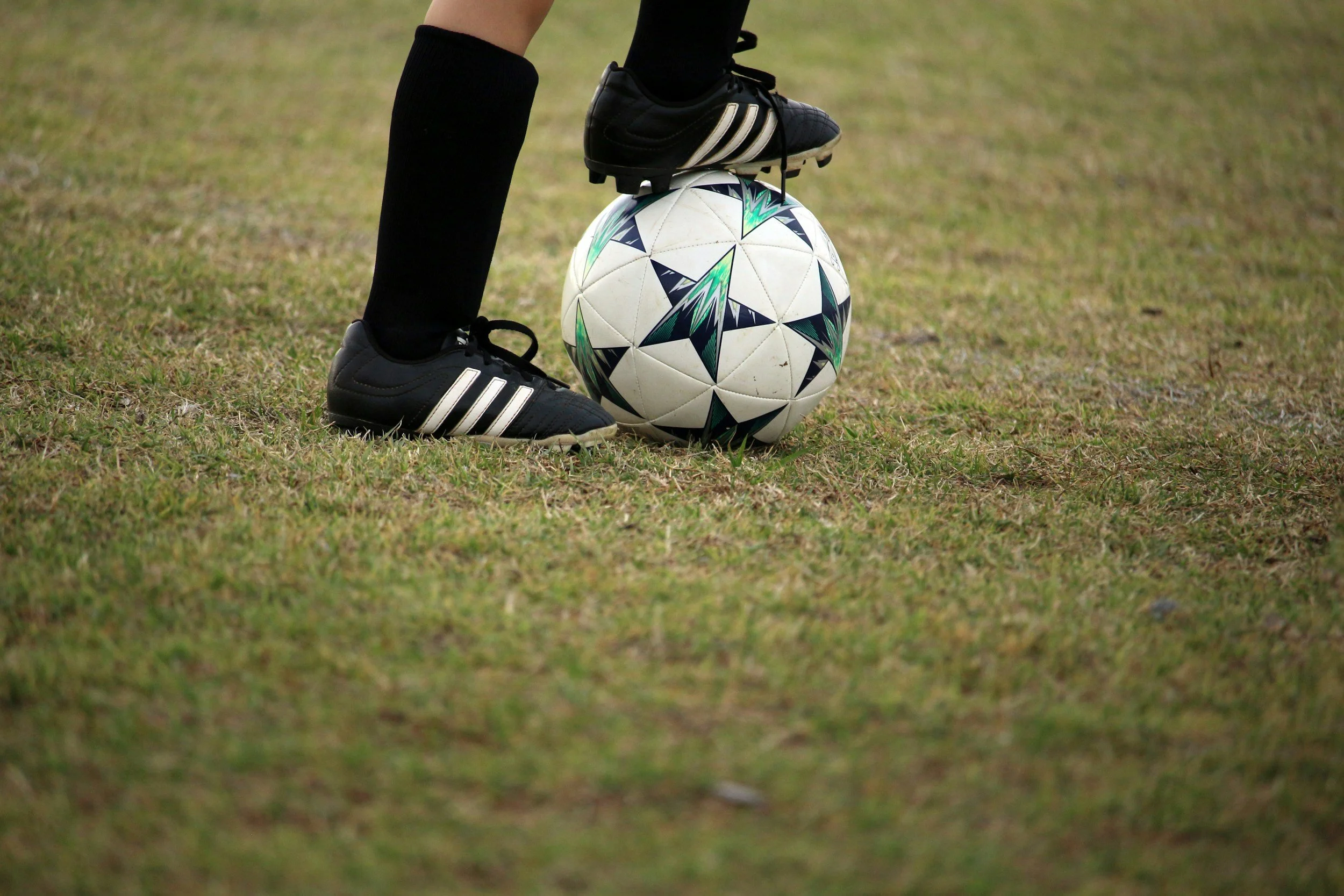 A person wearing black soccer cleats and black socks is standing on grass, with their foot on a white soccer ball decorated with green and black star patterns.