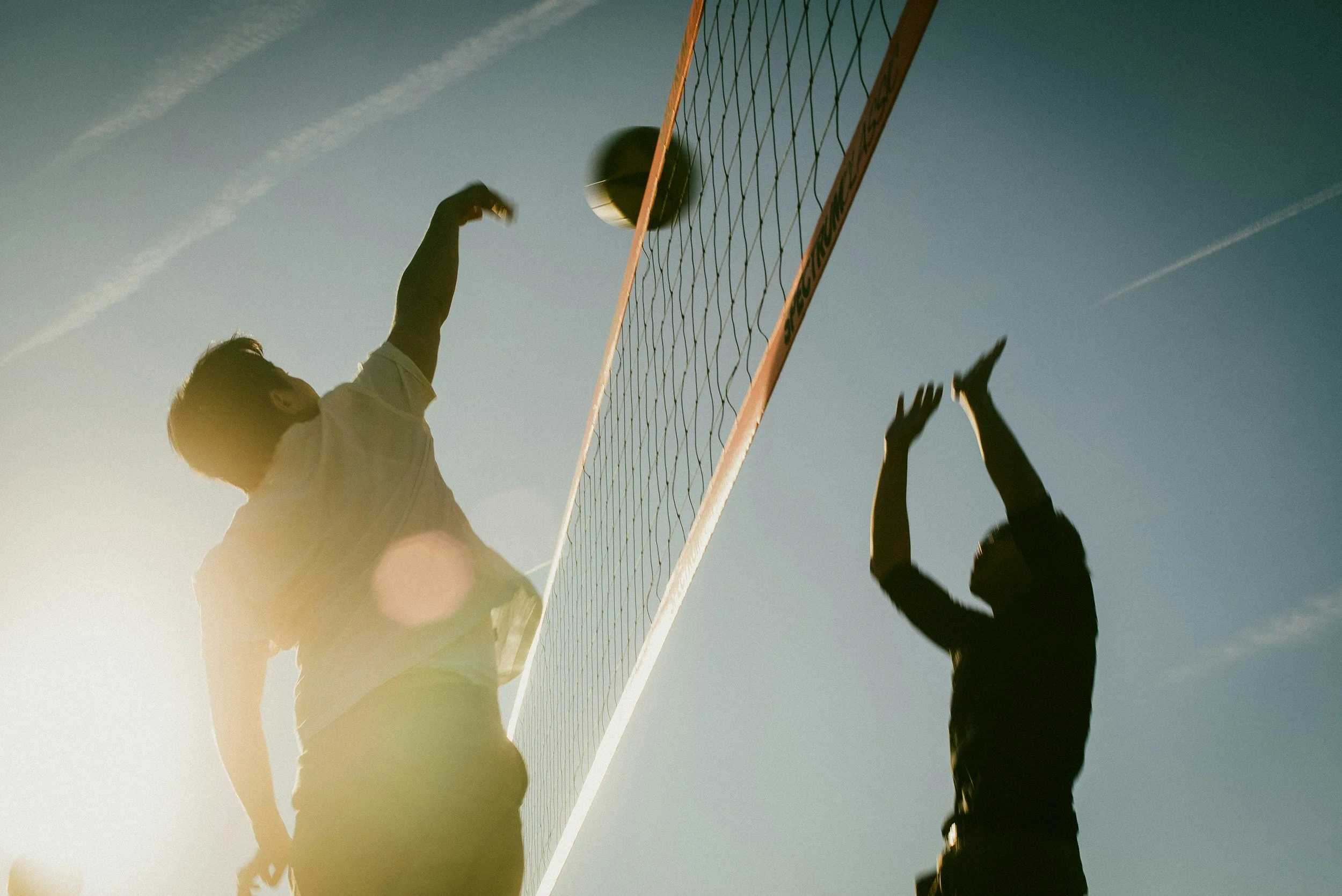Two people playing volleyball outdoors, one jumping to hit the ball over the net during daytime with a clear blue sky.
