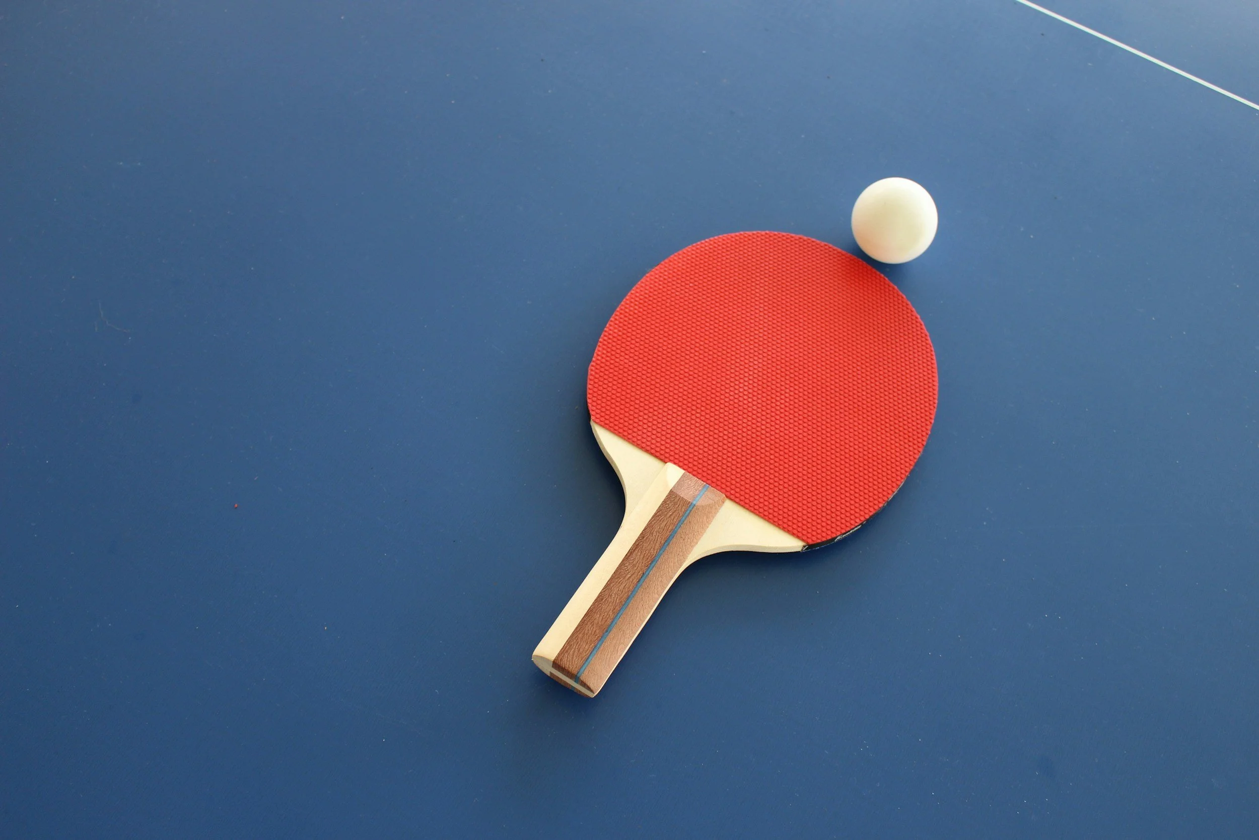 Two ping pong paddles, one with a red surface and one with a white surface, and a white ping pong ball on a blue table.