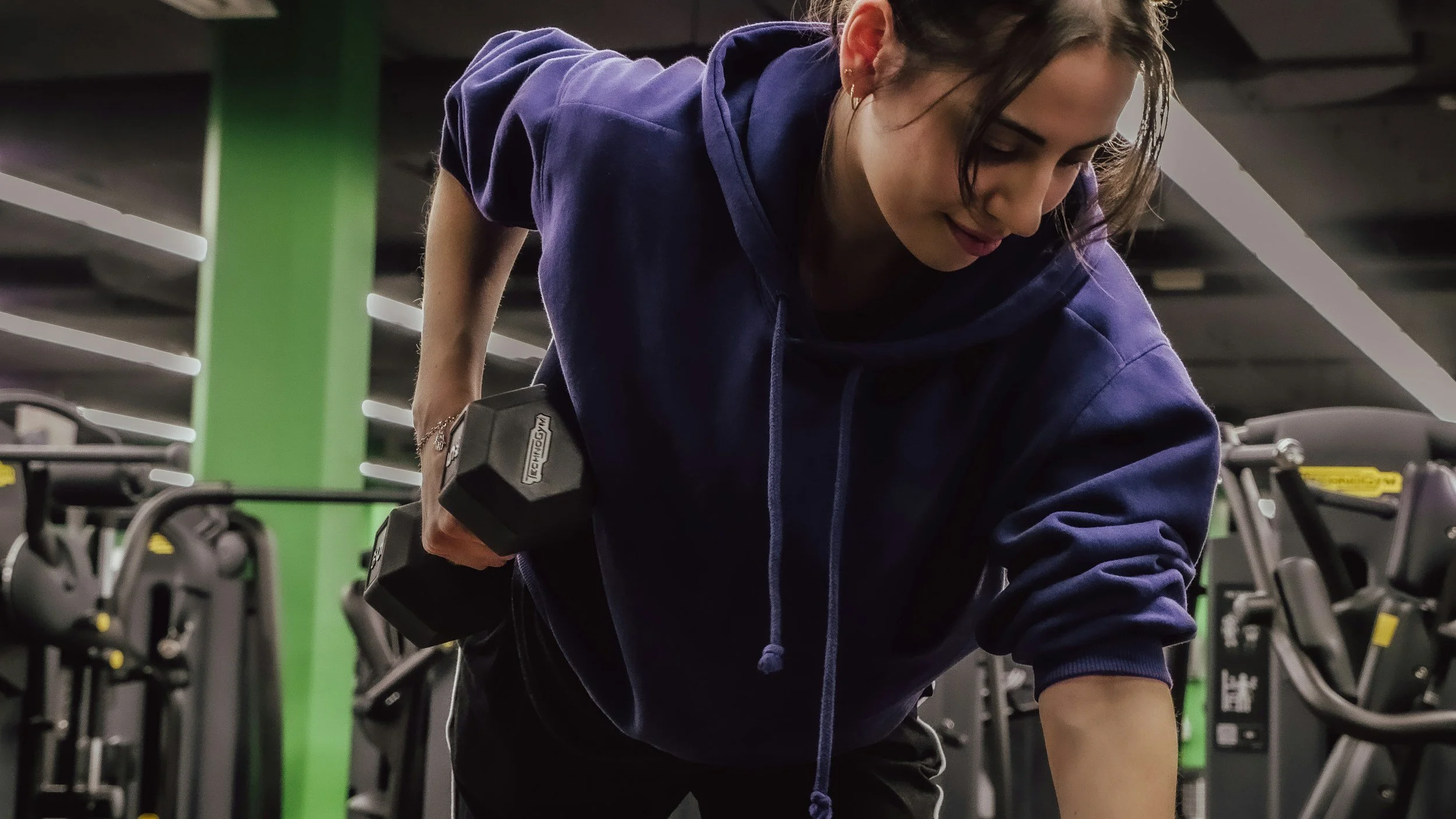 A woman in a purple hoodie lifting a dumbbell in a gym with green and black workout equipment in the background.