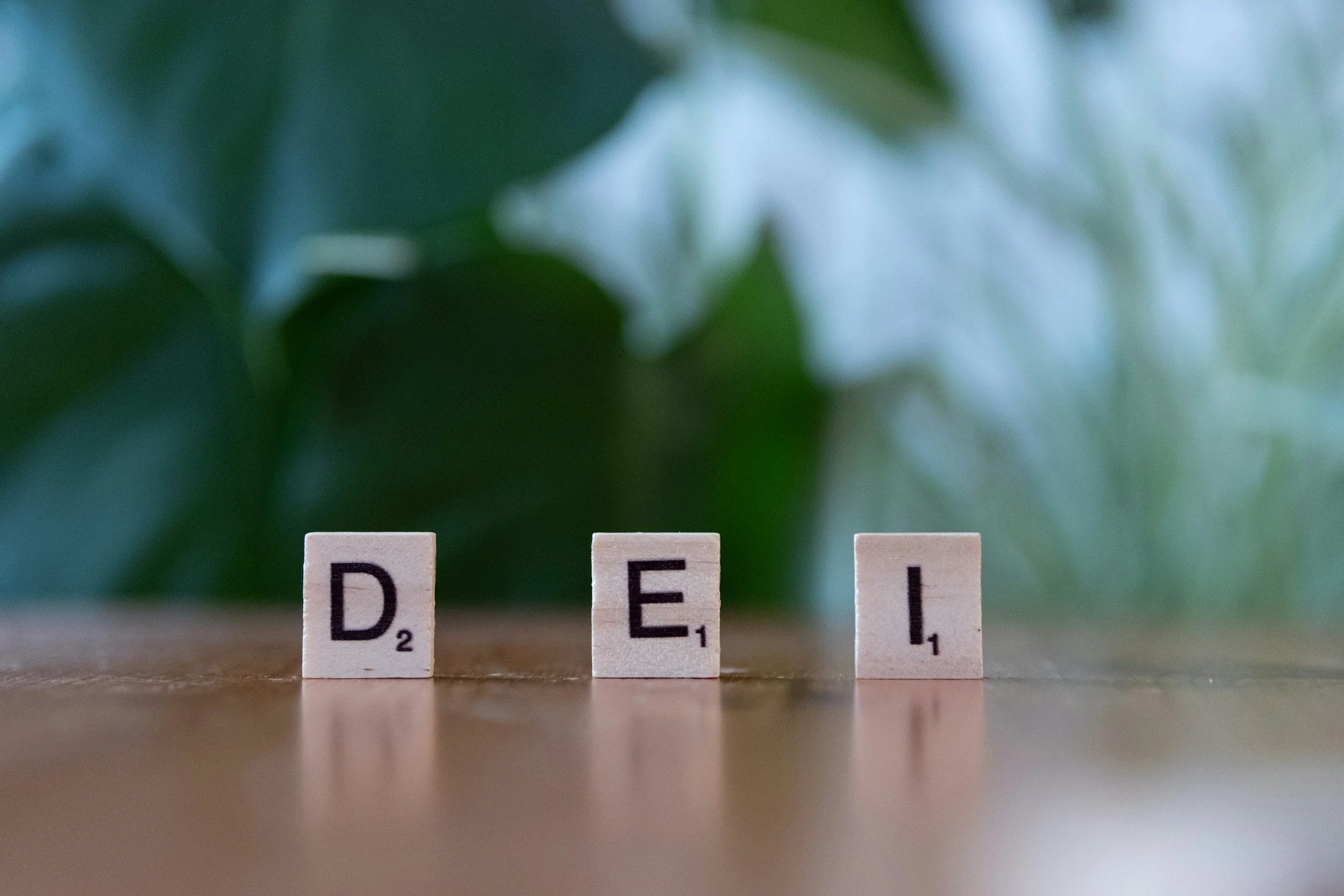 Close-up of three Scrabble letter tiles spelling the word 'DELI' on a wooden surface, with a blurred green leafy background.