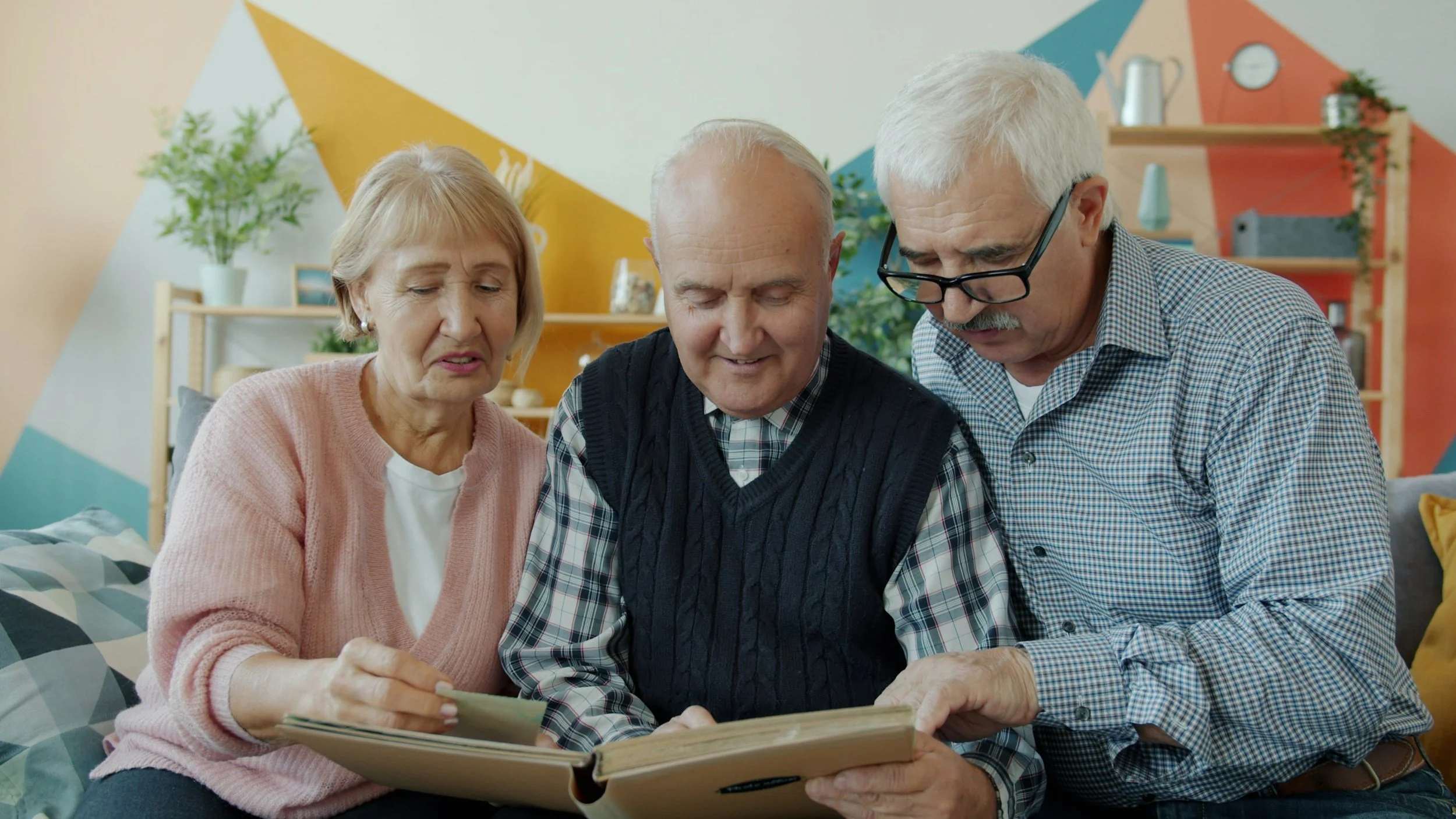 Three elderly adults, two men and one woman, sitting closely together on a bed, looking at a photo album or scrapbook, with a colorful geometric wall and plants in the background.