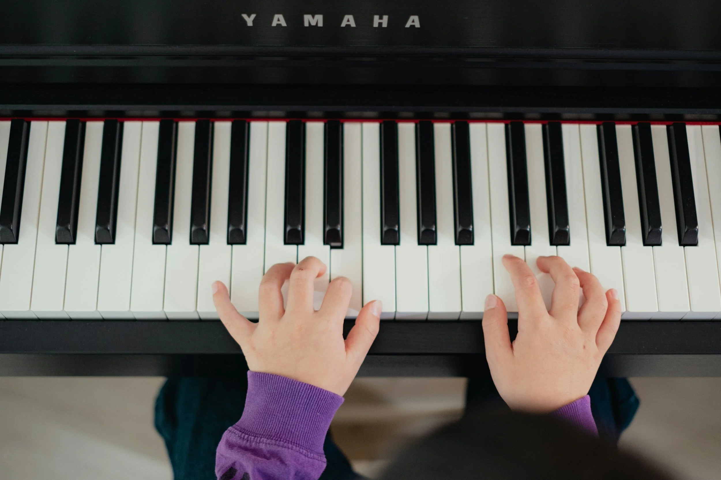 Top-down view of a digital piano with black and white keys, with two hands playing. The person is wearing a purple long-sleeve shirt, and only their hands and part of their arms are visible.