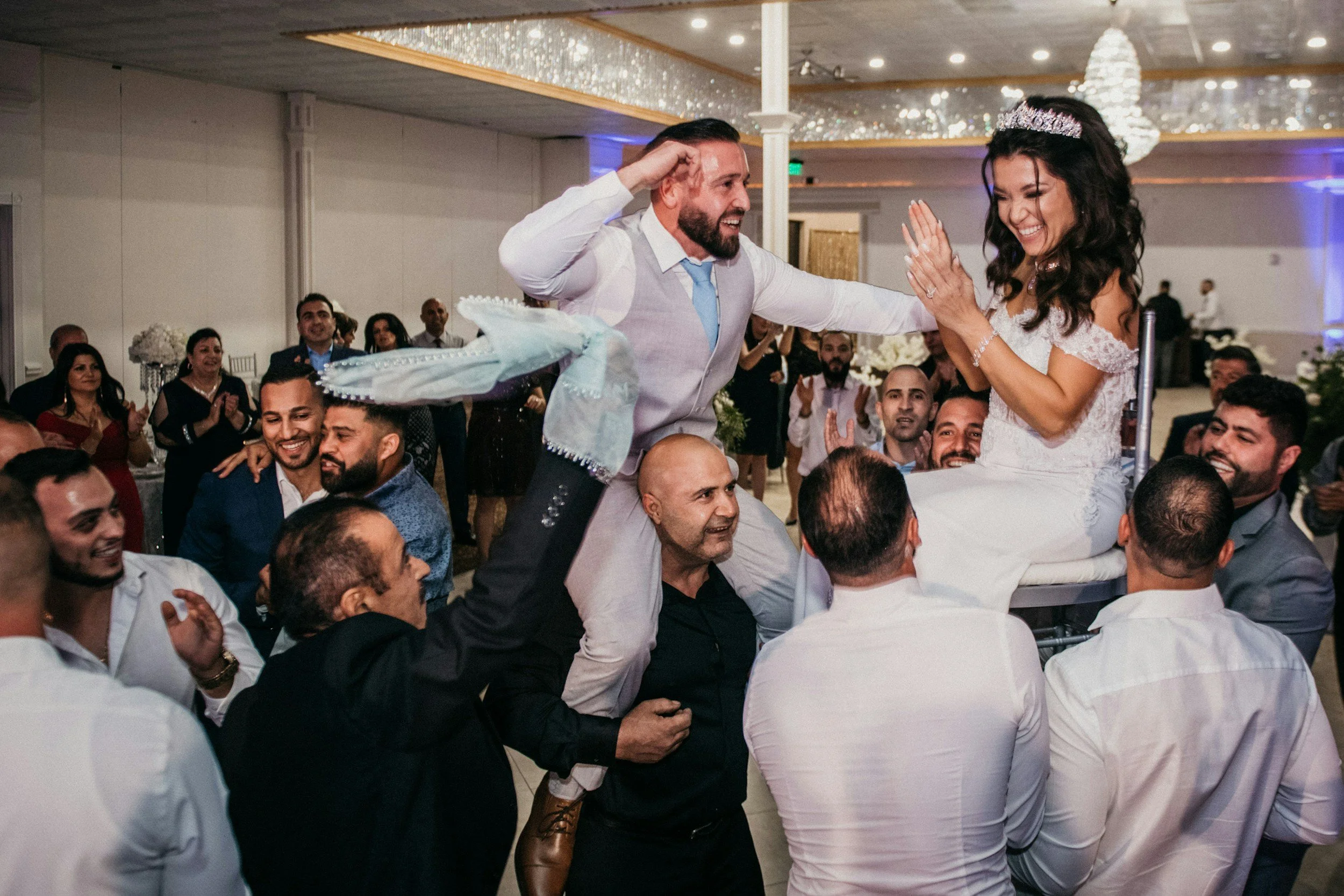 A wedding celebration with a bride in a white wedding dress and a tiara, and a groom in a gray vest and tie, dancing with guests in an indoor venue decorated with chandeliers and flowers.
