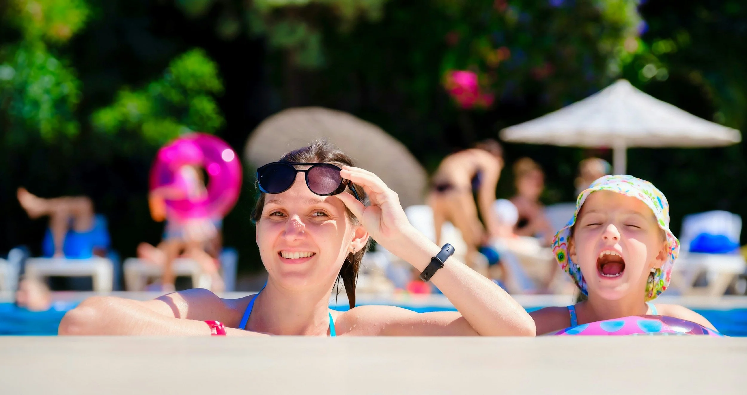 A woman and a young girl in a swimming pool, smiling and enjoying a sunny day, with trees and poolside umbrellas in the background.