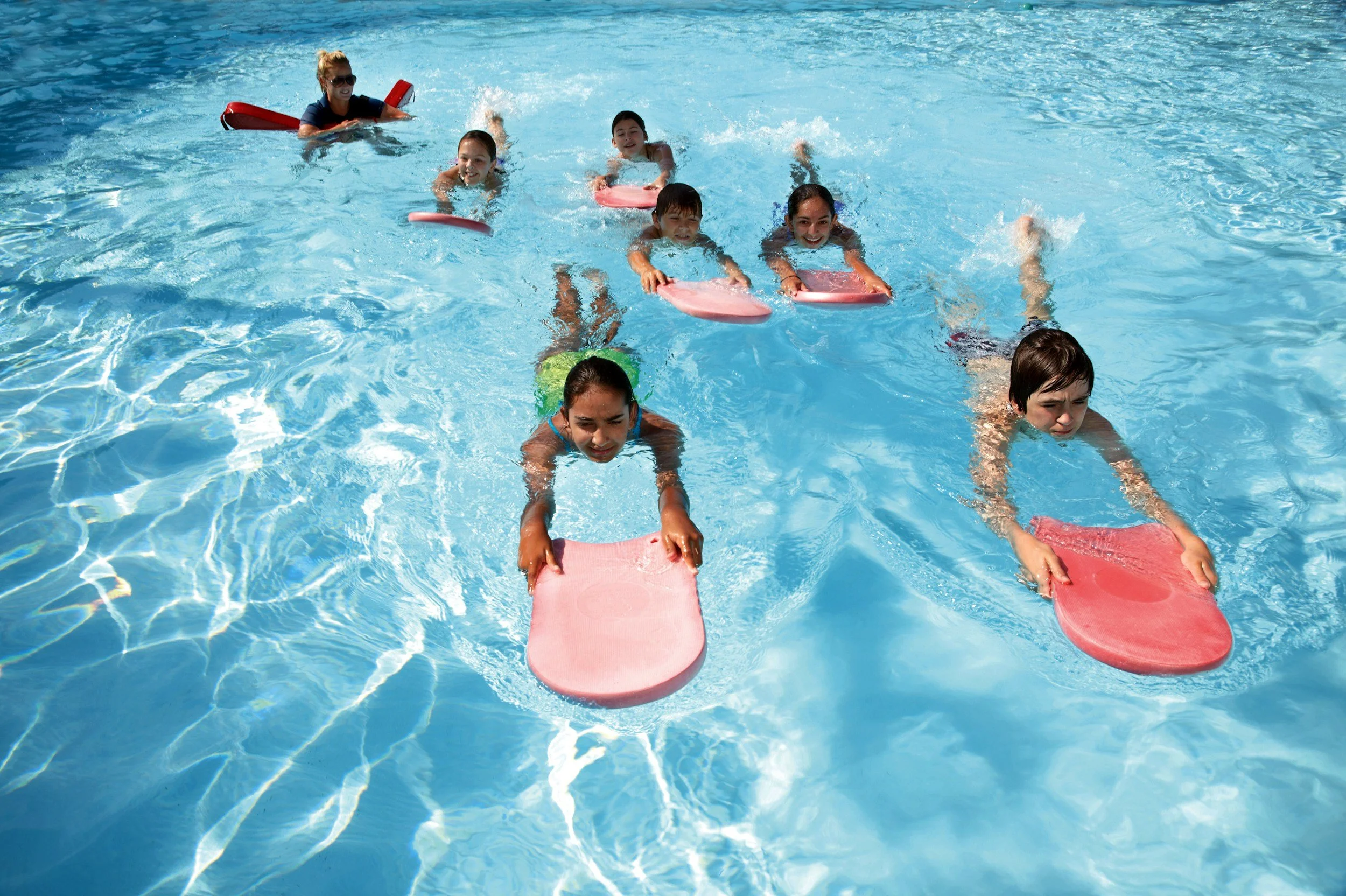Group of children and an adult swimming and floating on pink kickboards in a bright blue swimming pool.