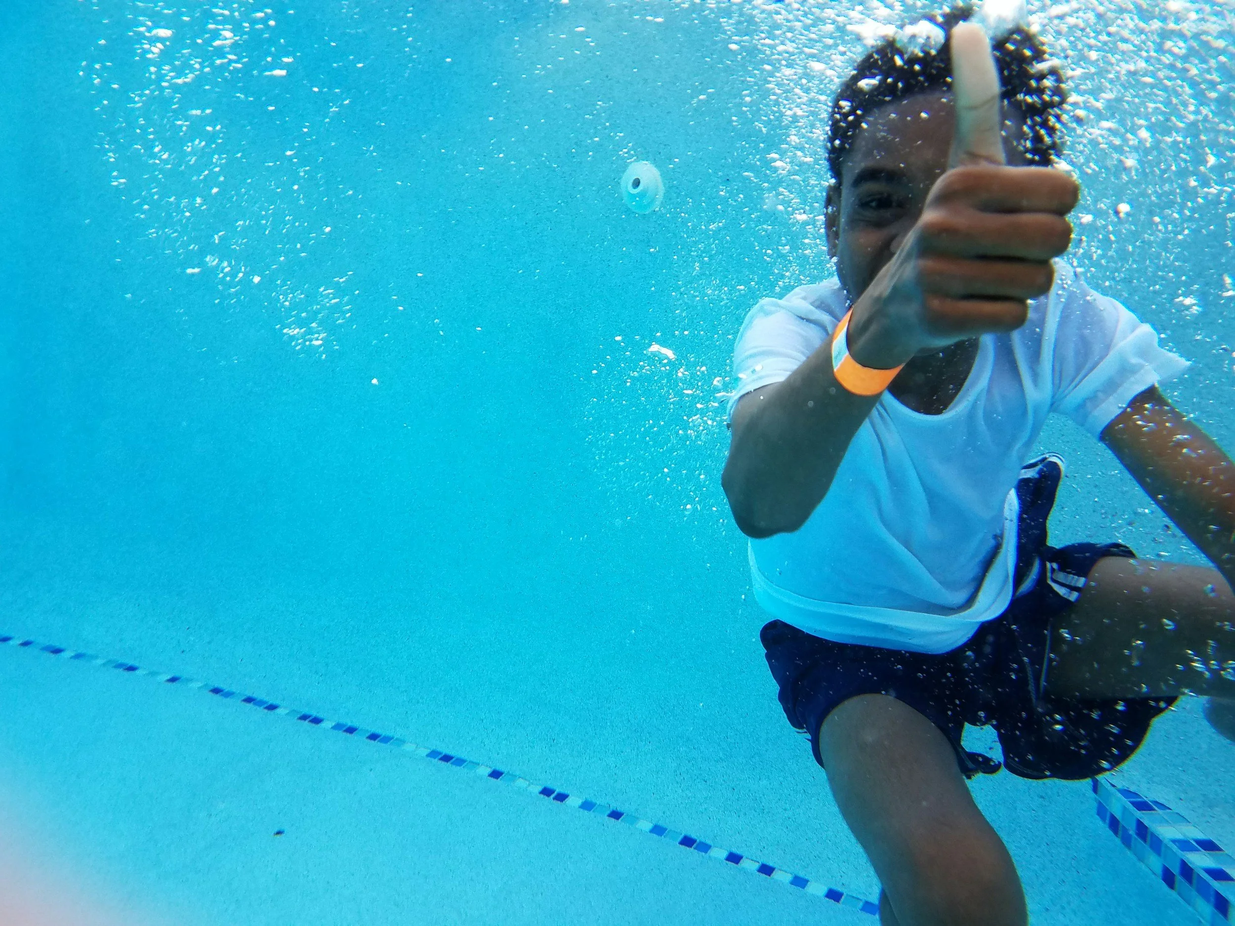 A person in a white shirt giving a thumbs-up underwater in a swimming pool.