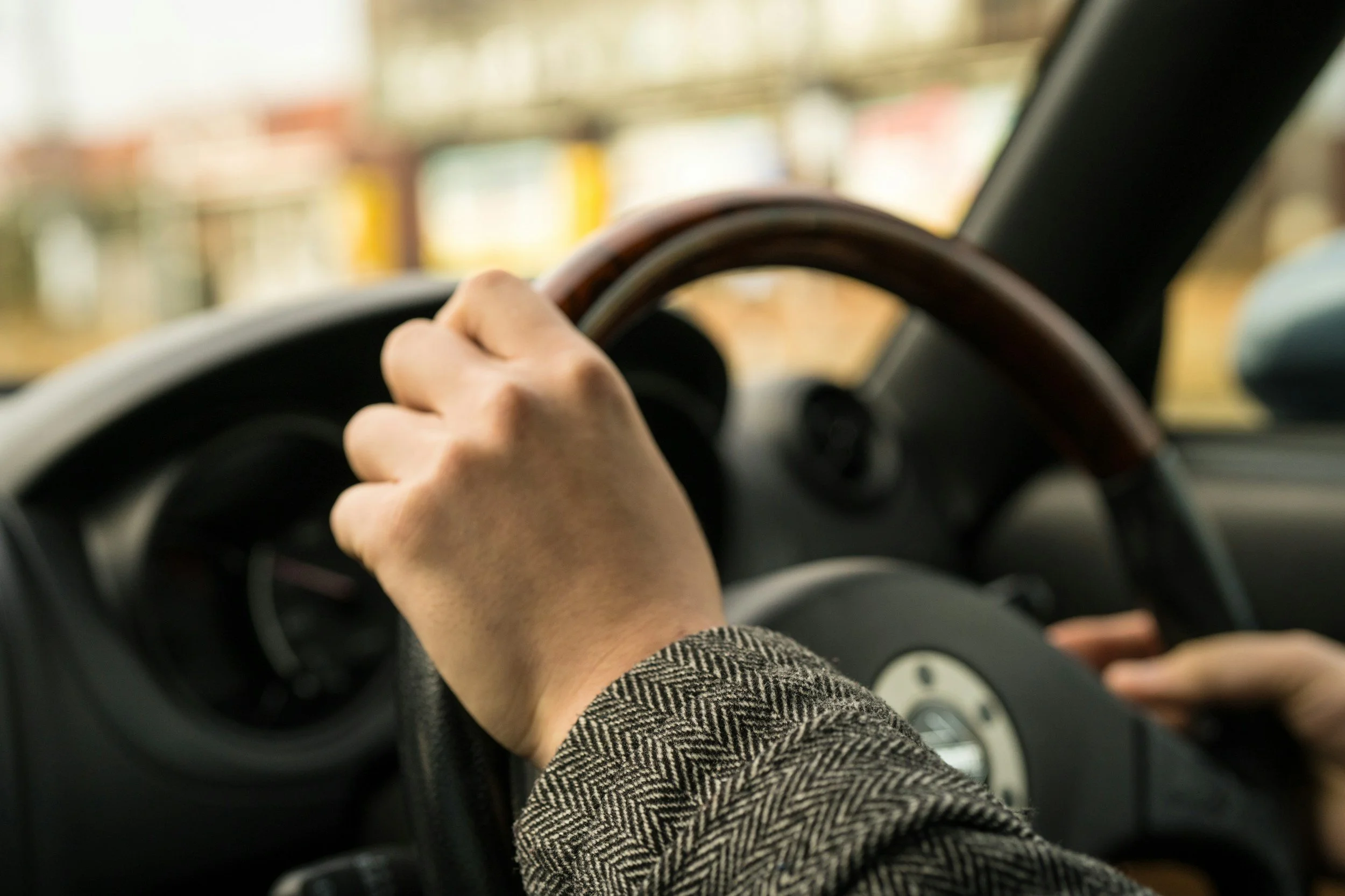 A person driving a car, holding the steering wheel with their left hand, wearing a checkered blazer.