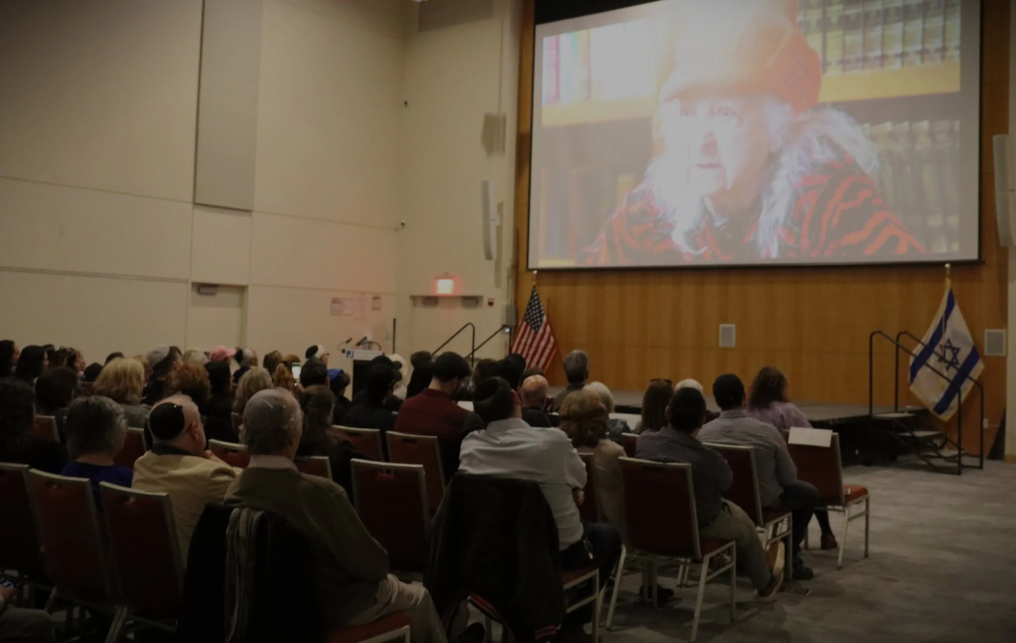 Audience seated in a conference room watching a presentation on a large screen. The screen displays a close-up image of an elderly woman with grey hair, a orange knit hat, and a red and black patterned shirt, with a background of bookshelves filled with books. The room has a wooden-paneled wall and flags, including an American flag and another flag, near the stage.