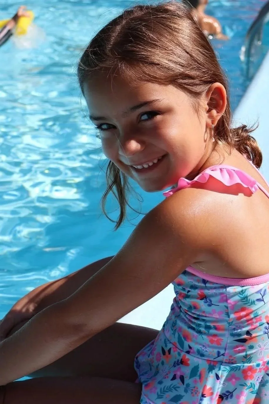 Young girl with brown hair smiling at swimming pool, wearing a colorful floral swimsuit with ruffled pink straps.