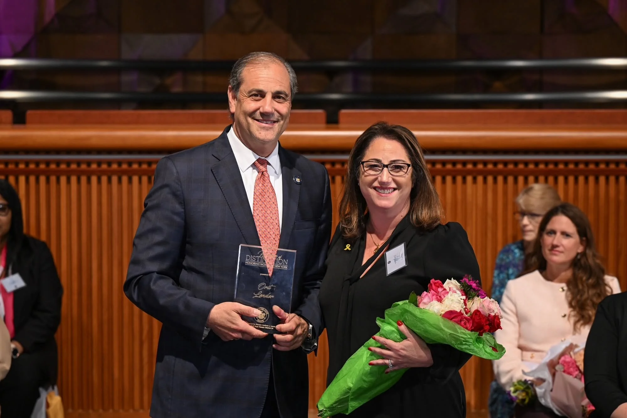 A man in a dark suit and a woman in glasses and a black outfit are smiling at an award ceremony. The woman is holding a bouquet of pink and white flowers, and the man is holding a clear plaque or trophy. Several people can be seen seated in the background.