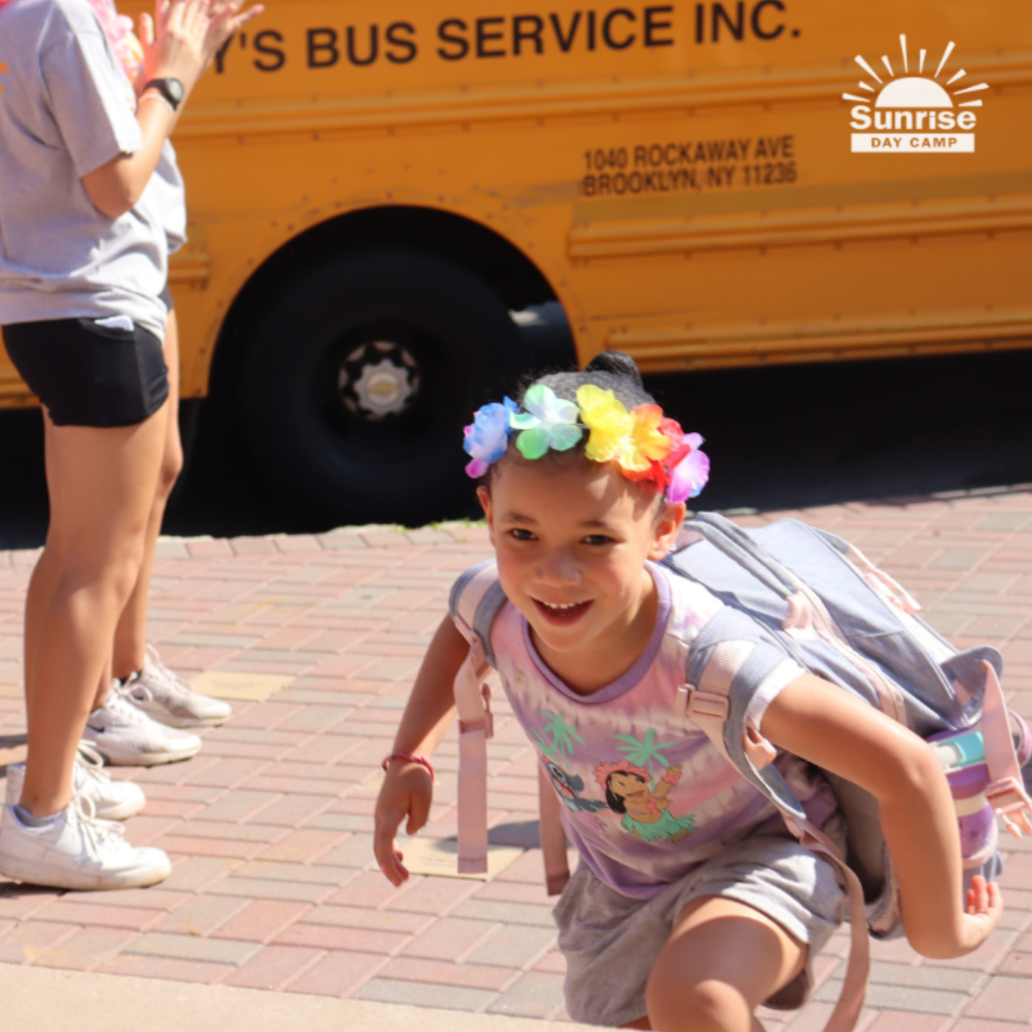 A young girl with a colorful flower crown, smiling and crawling on a brick sidewalk, near a yellow school bus.