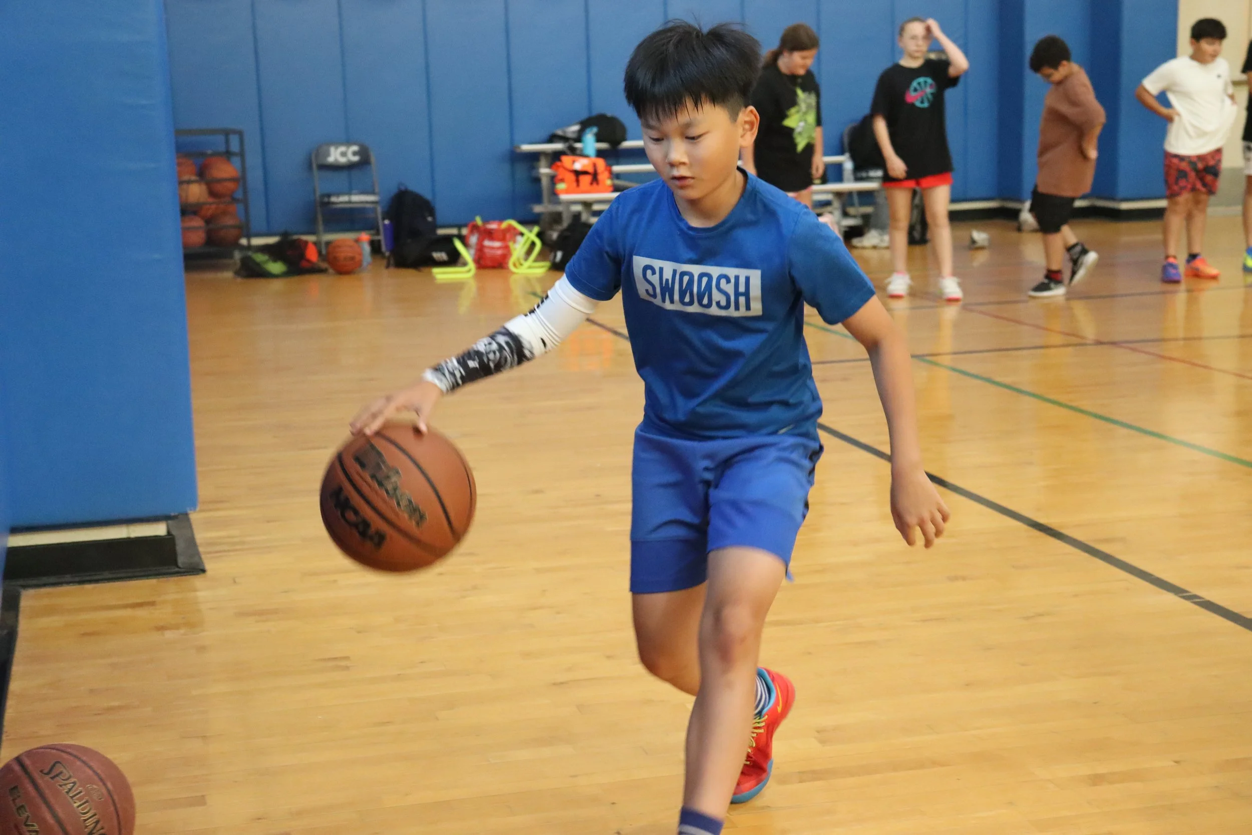 A young boy in a blue shirt and shorts dribbling a basketball on an indoor basketball court, with other children and sports equipment visible in the background.