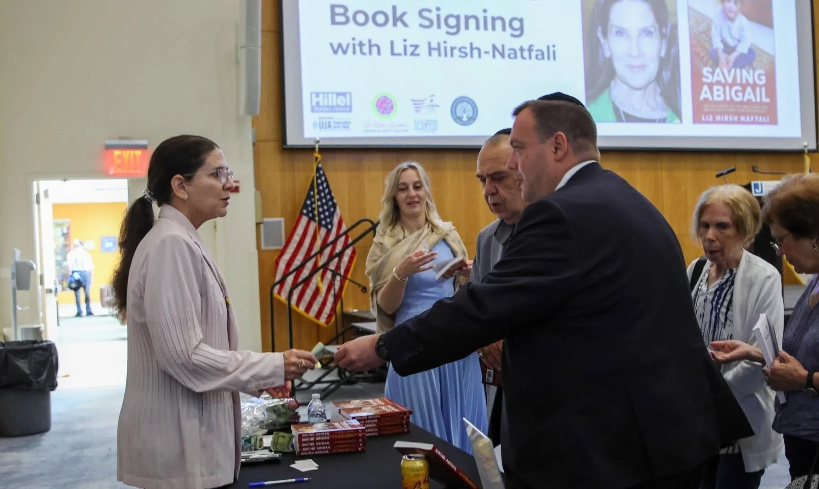A book signing event with Liz Hirsh-Naftali, featuring a woman in a pale pink suit handing a book to a man in a dark suit, with other women and an American flag in the background.