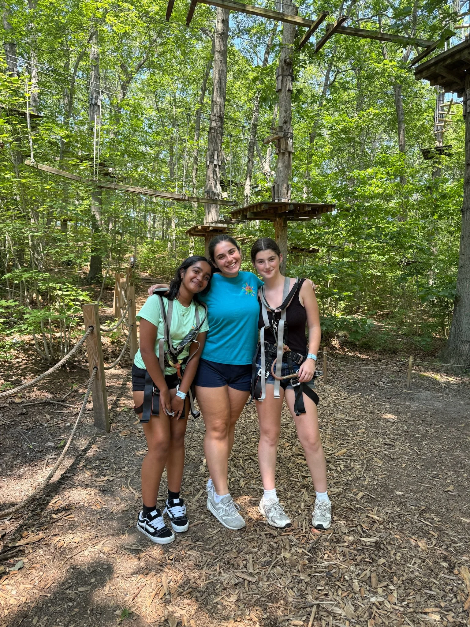 Three young women standing together in a forest, smiling. They are wearing casual clothing with harnesses for a ropes course or zip-lining adventure, with trees and wooden platforms in the background.
