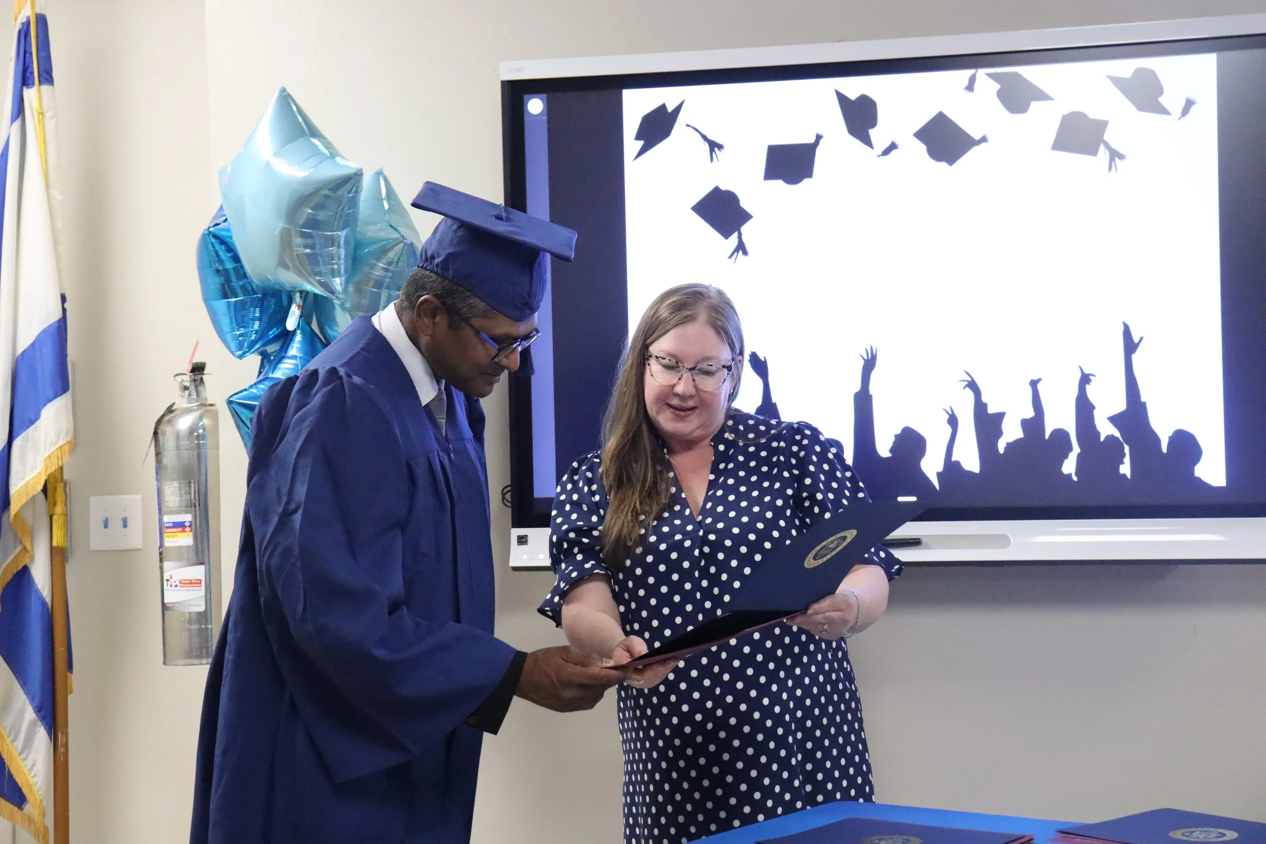 A man in a graduation gown and cap is standing next to a woman in a polka-dot dress, both looking at a vinyl record she is holding. Blue balloons are attached to a fire extinguisher on the side, and a screen with black silhouettes of graduation caps and hands in the air is in the background.