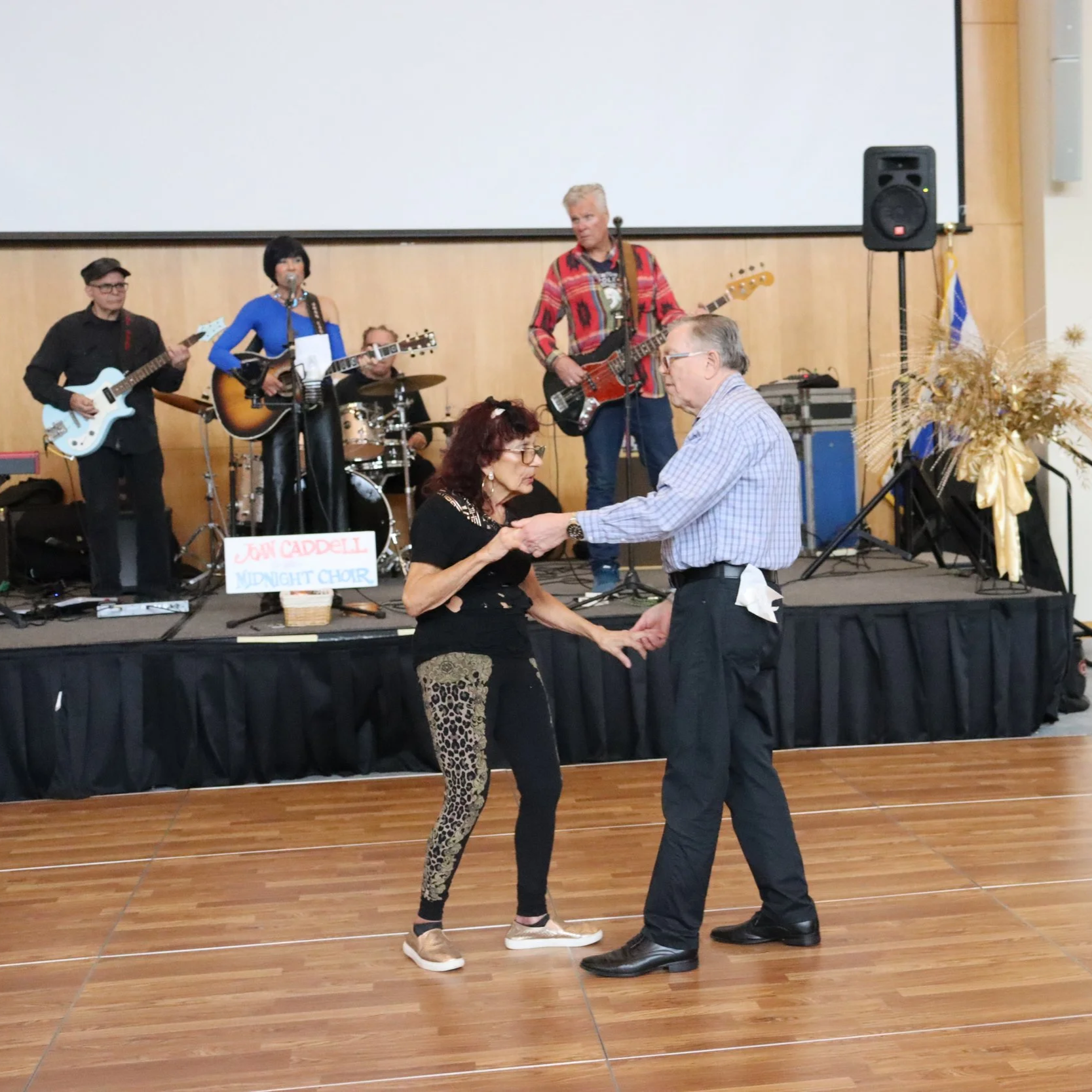 Couple dancing in front of a band playing music on a stage at an indoor event, with a woman and a man holding hands on the wooden dance floor.