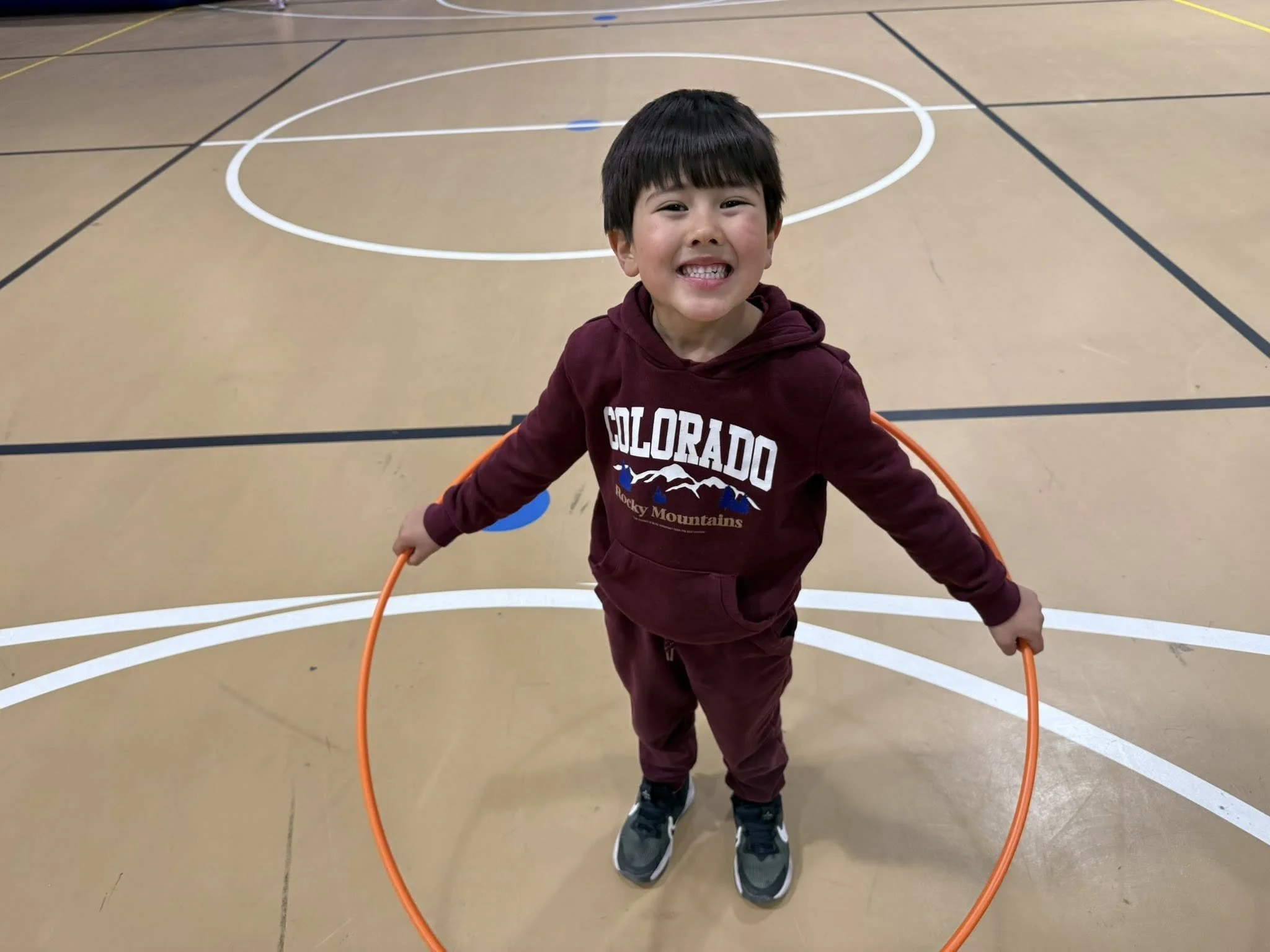 Young boy smiling and jumping inside an orange hula hoop on a gymnasium floor with black and white court lines.