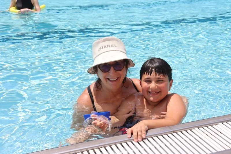 A woman and a young boy smiling while swimming in a pool, the woman wearing a white hat with 'Chanel' written on it and sunglasses.